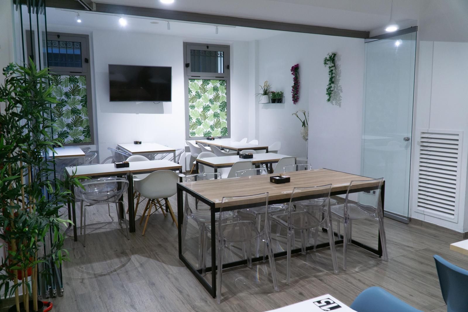 Una zona salotto interna moderna e luminosa, con tavoli in legno chiaro, sedie trasparenti e pareti bianche decorate con piante.