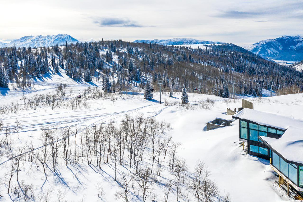 An aerial view of a house in the middle of a snowy field with mountains in the background.