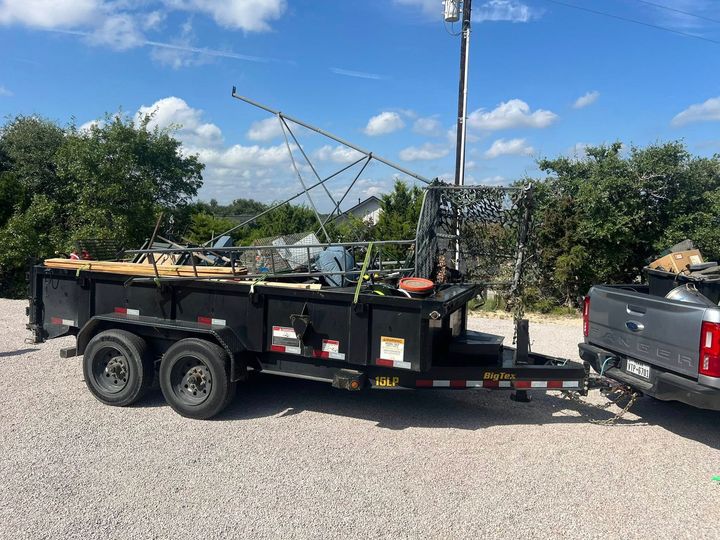 A dark dump trailer loaded with construction equipment and metal beams, hitched to a gray pickup truck on a gravel lot.