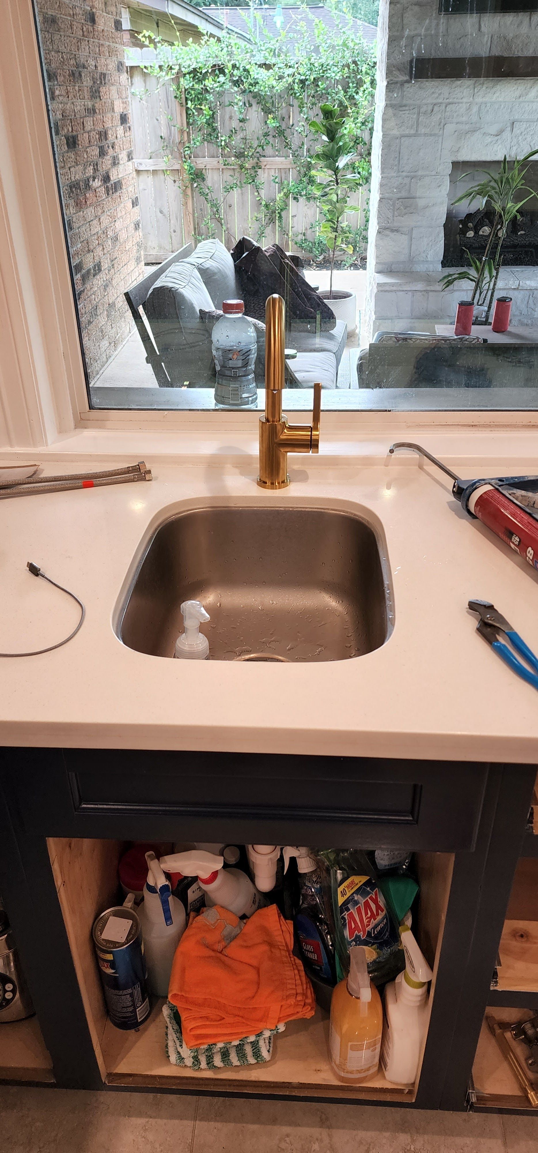 White countertop over a dark cabinet filled with cleaning supplies. Window in the background.