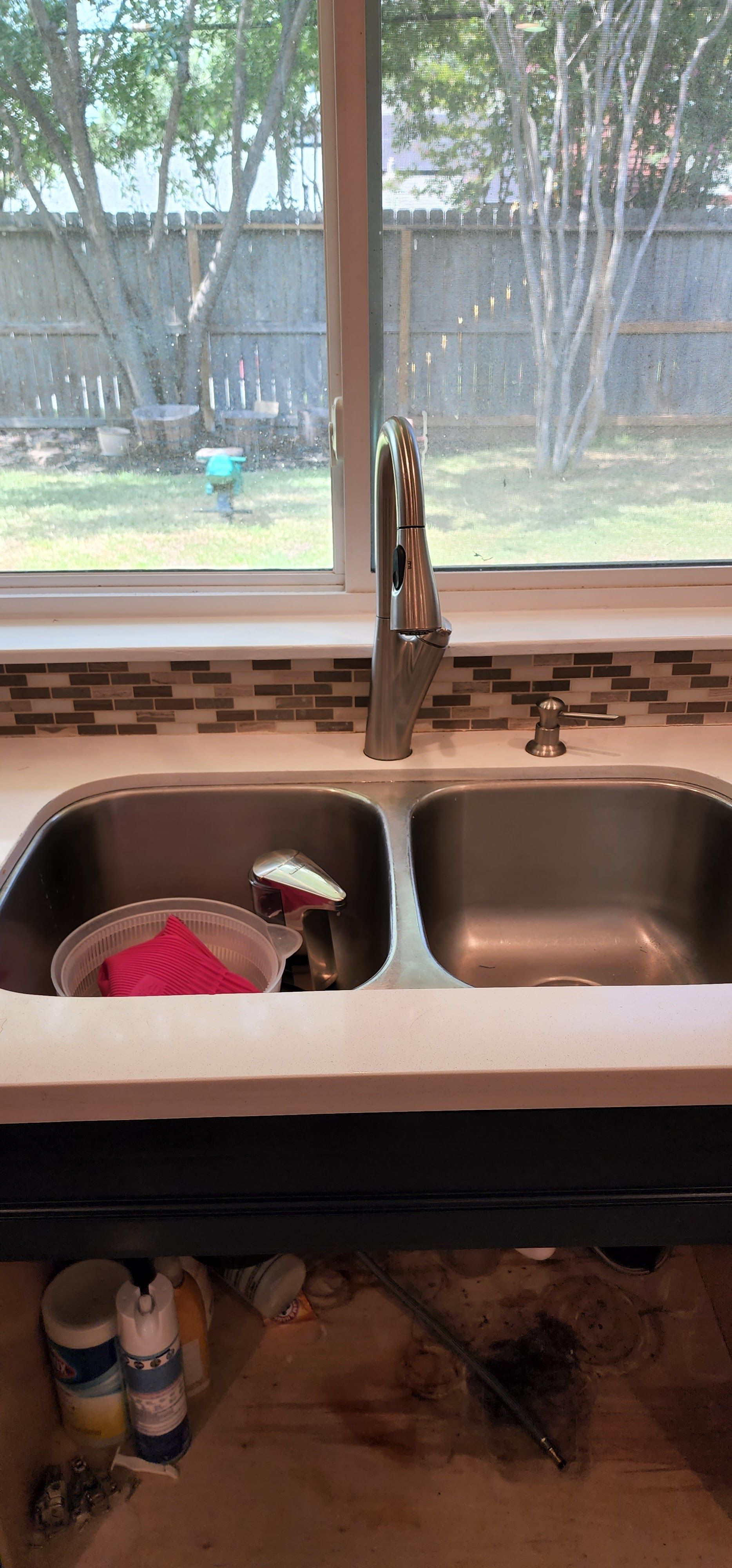 Kitchen sink with a window view of a yard and trees. A dish rack with pink sponge and spray bottle.