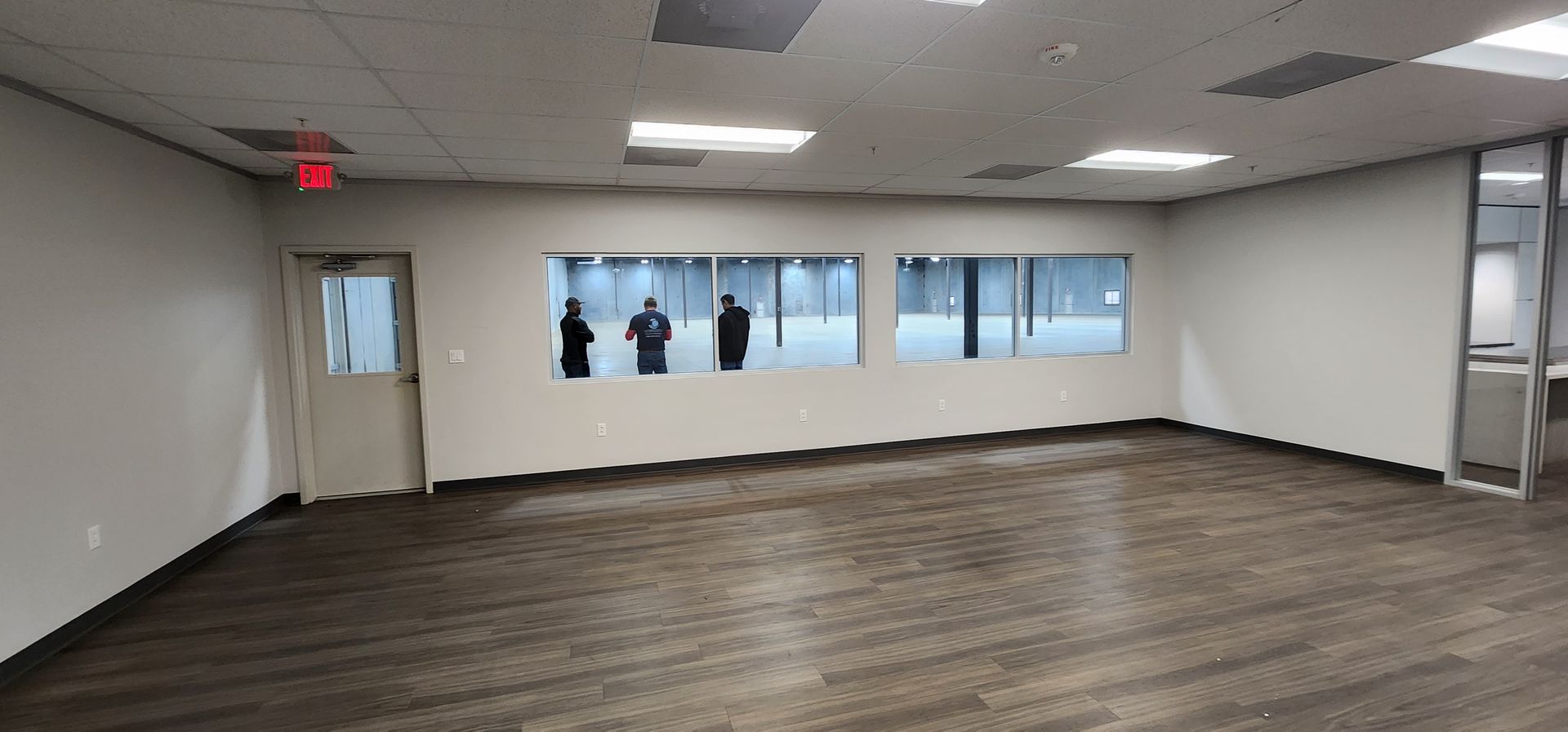 An empty room with three figures visible through windows. Beige walls, brown carpet, ceiling lights.