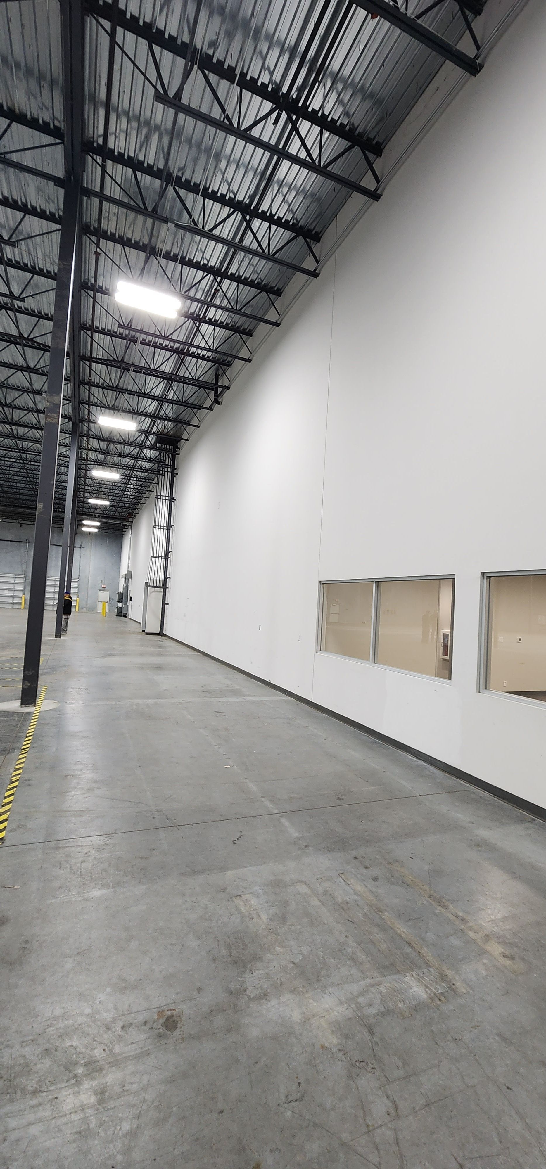 Warehouse interior with high ceiling and concrete floor. White wall on the right.  Metal beams overhead and a row of lights.