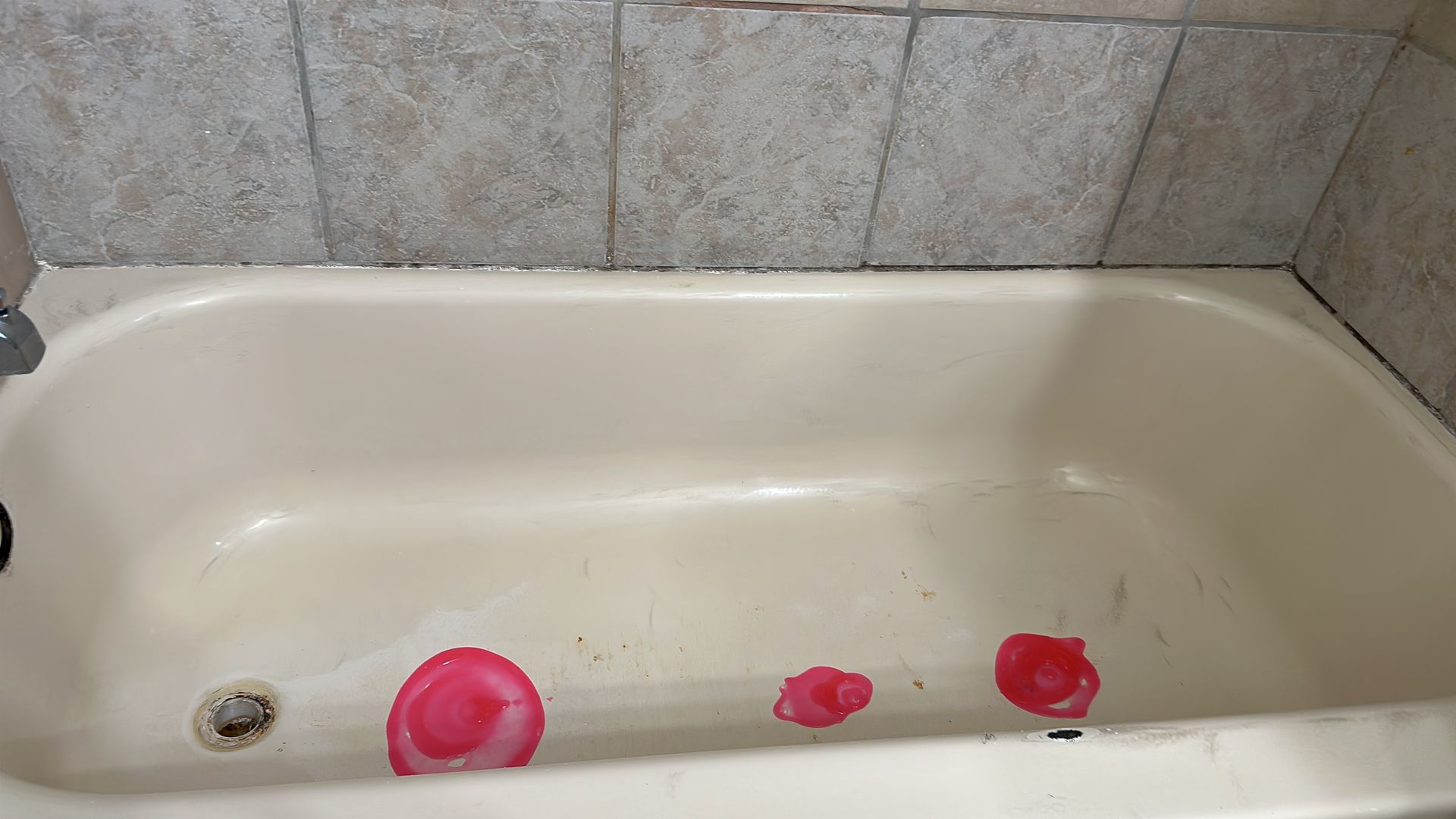 A dirty bathtub with three red bath bombs in it, under beige tiles.