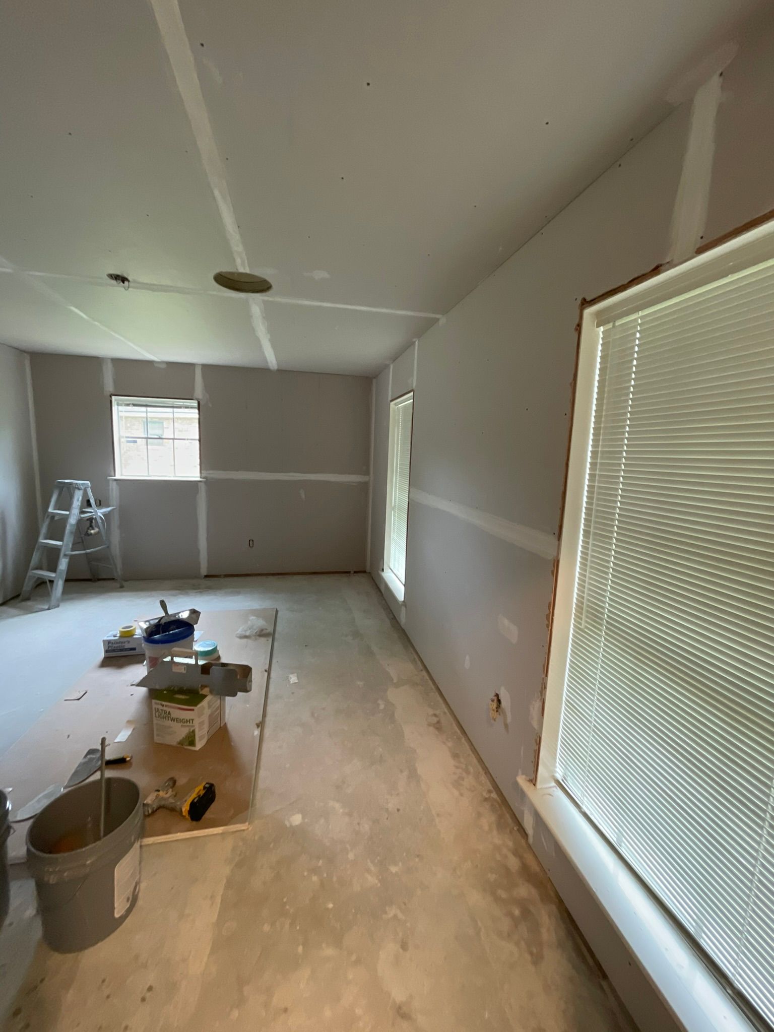 Room under construction, drywall installed. Window with blinds, ladder, tools, and buckets are visible.
