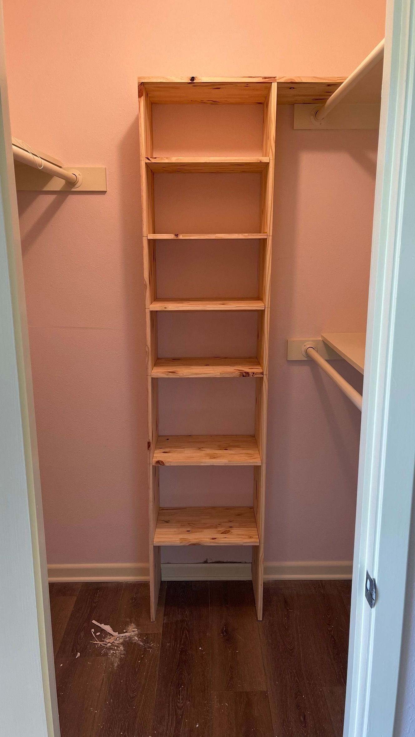 A tall, wooden shelf unit inside a closet, against a pink wall and dark brown floor.