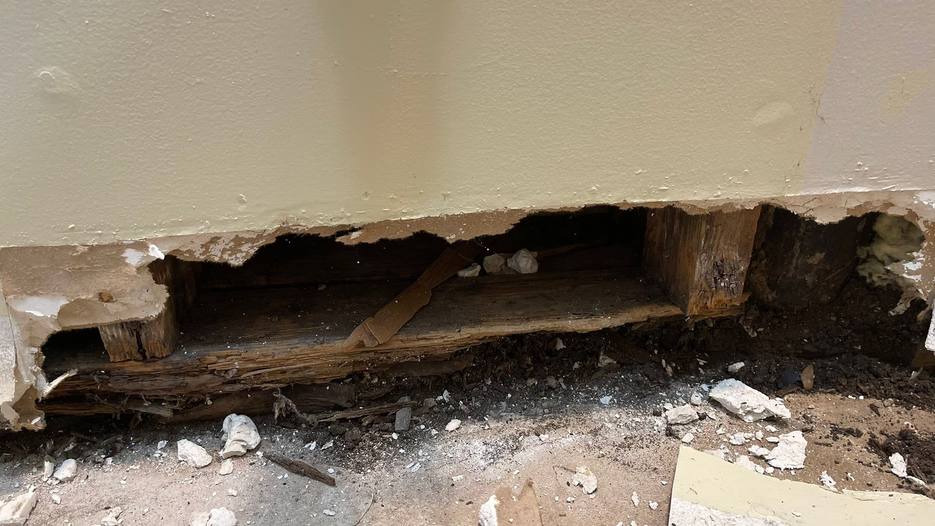 Hole in wall exposing wooden structure, with crumbling plaster and debris on floor.