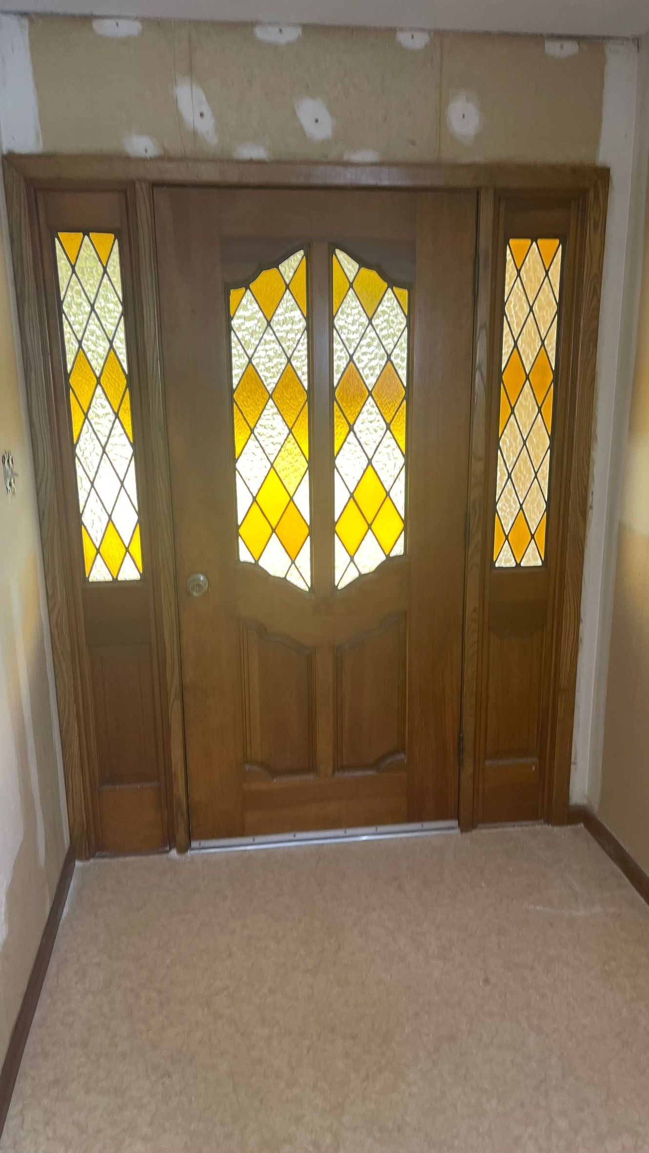 Wooden entry door with stained glass panes, flanked by side windows, beige carpet.