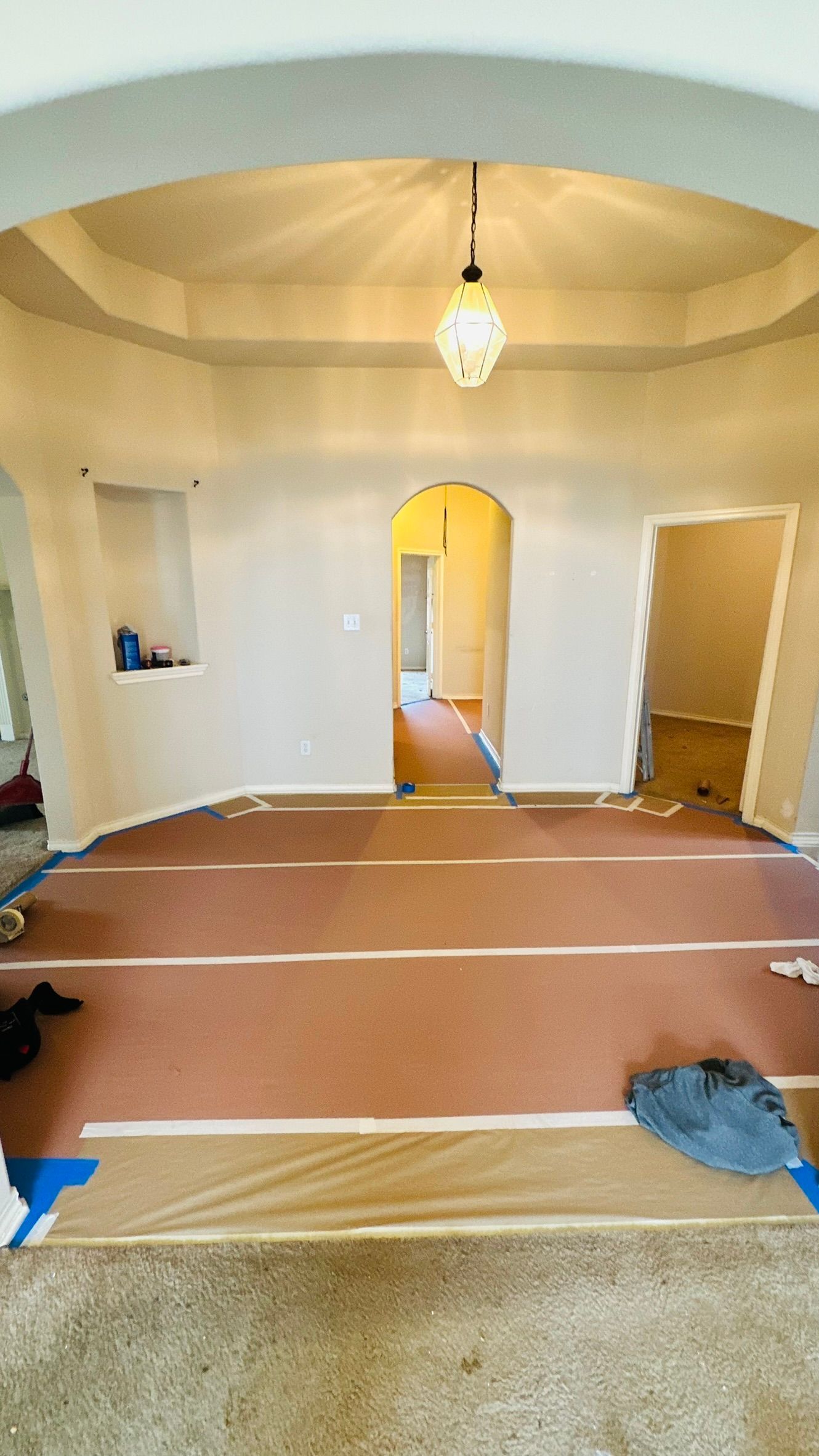 Interior room being renovated with protective floor covering. Neutral walls, arched doorways, and a hanging light.
