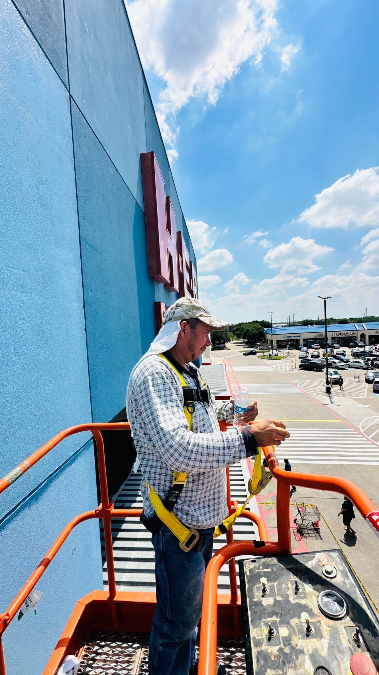 Man in lift bucket working on building signage; blue facade, bright sky, parking lot view.