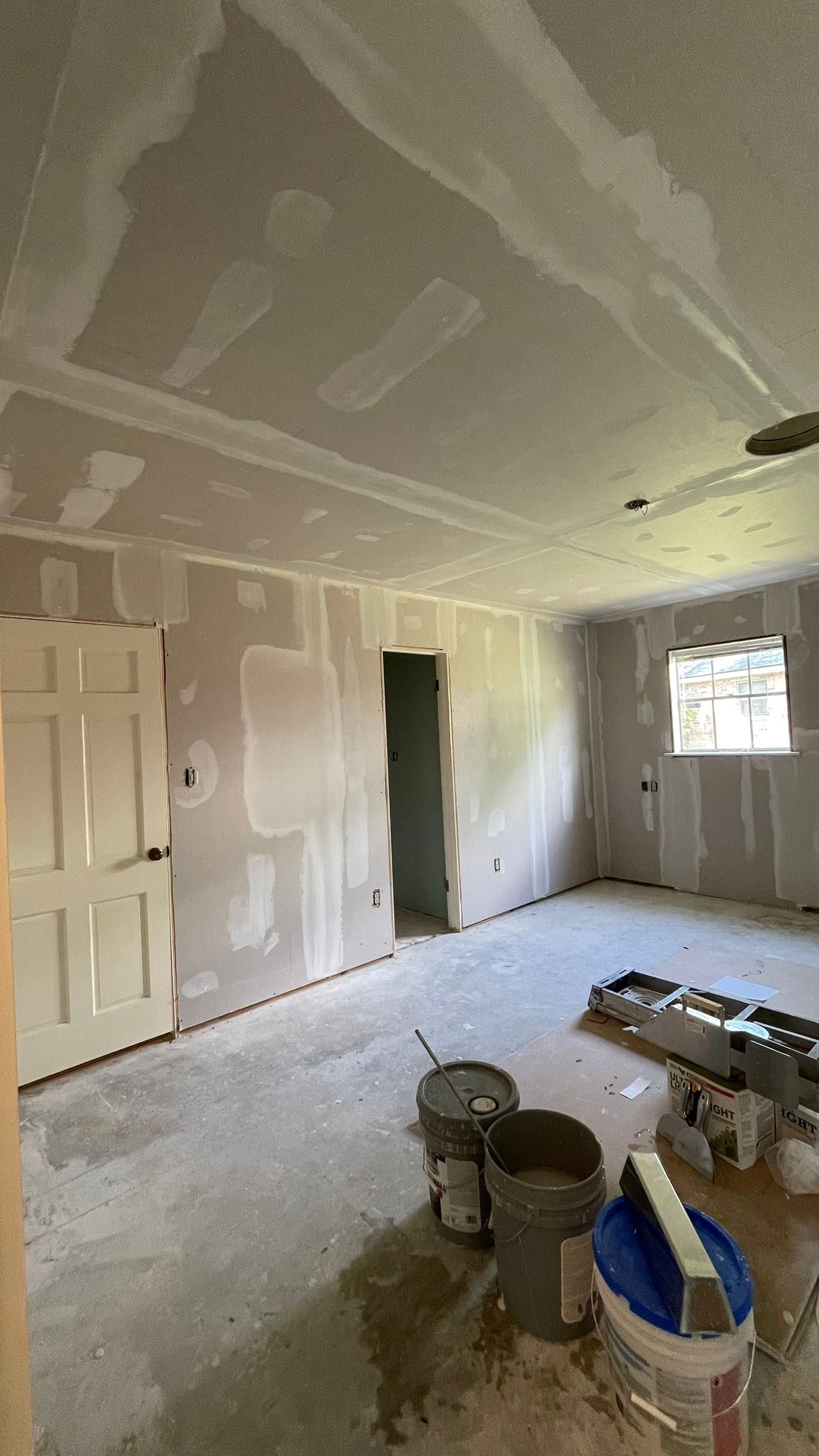 Interior of a room under construction, with drywall on walls and ceiling, buckets, and a door.