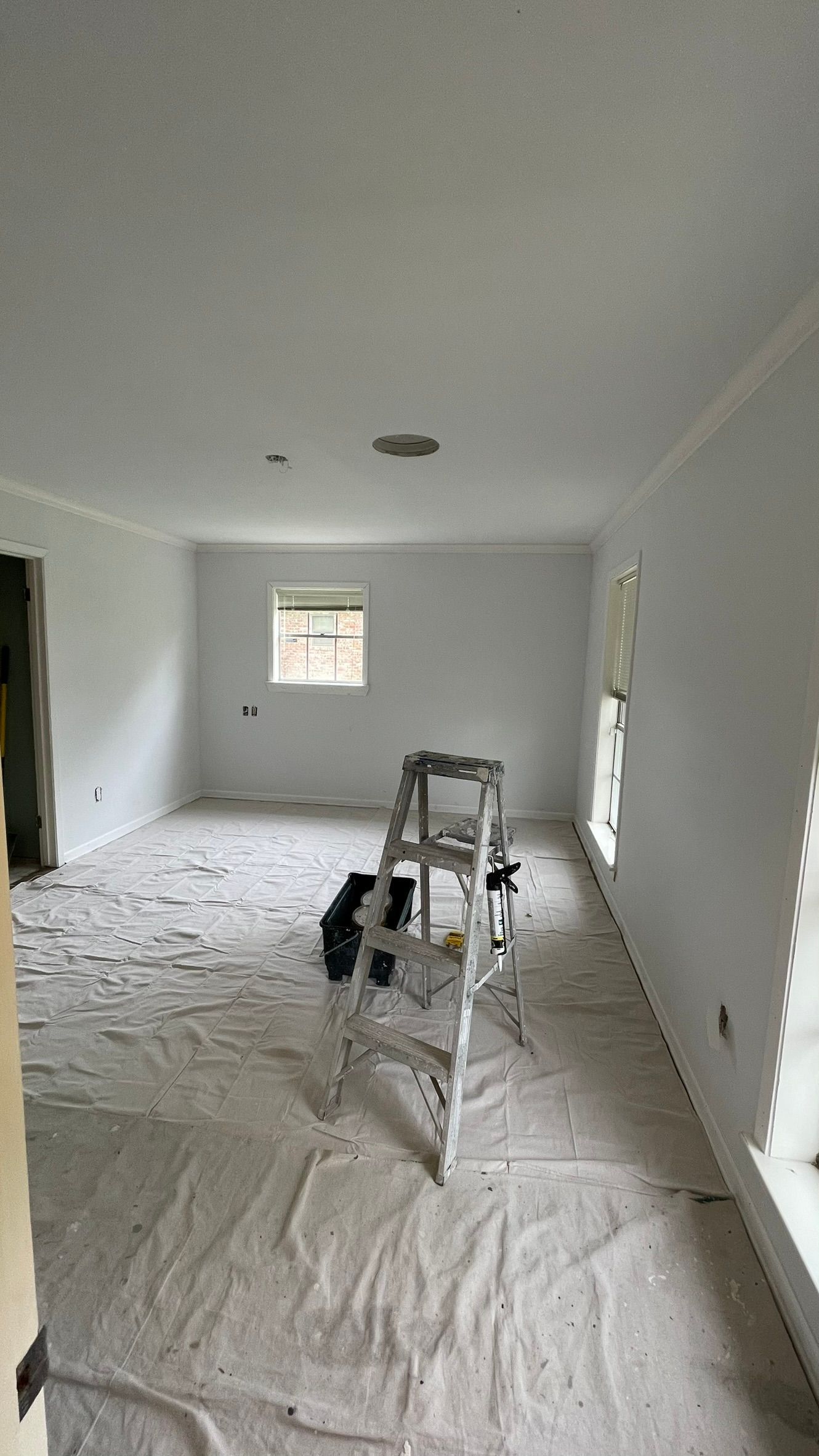 Room being painted; drop cloths on floor, ladder, white walls and ceiling, window, doorway.