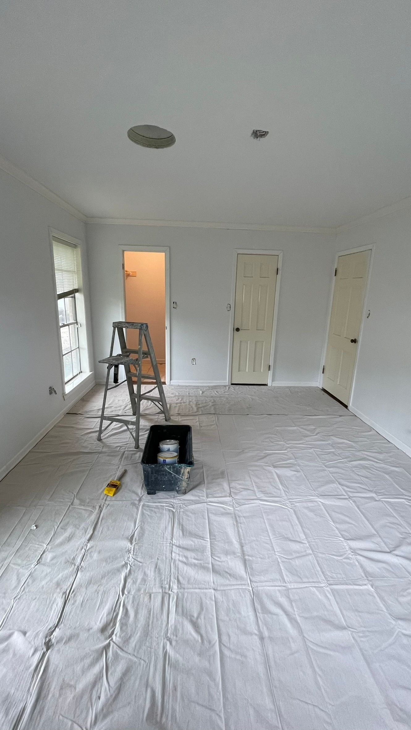 Room with white walls, doors, and ceiling, covered floor, ladder, and paint supplies.