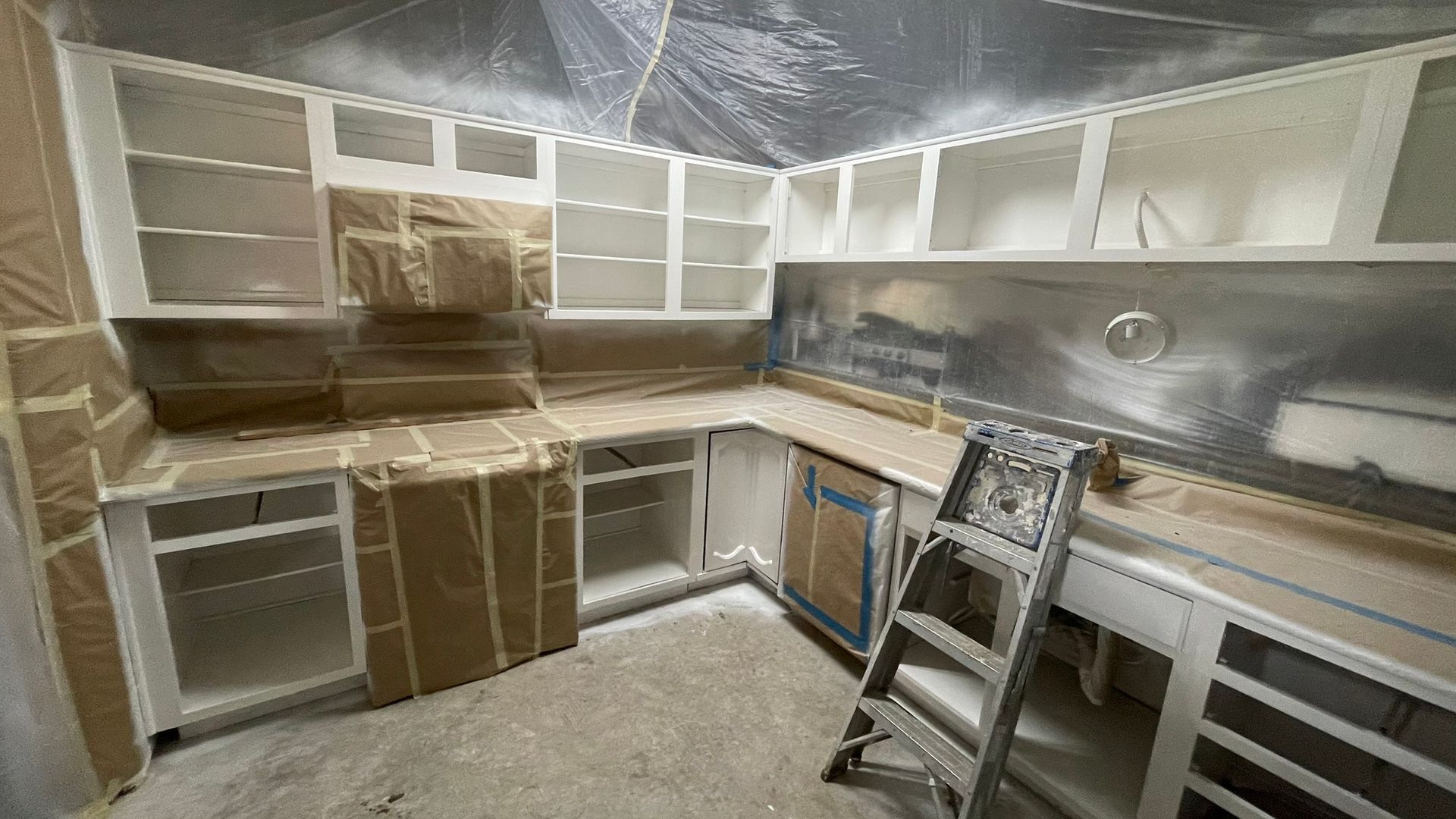 Kitchen cabinets being painted white, masked with brown paper and plastic sheeting. A stepladder stands nearby.