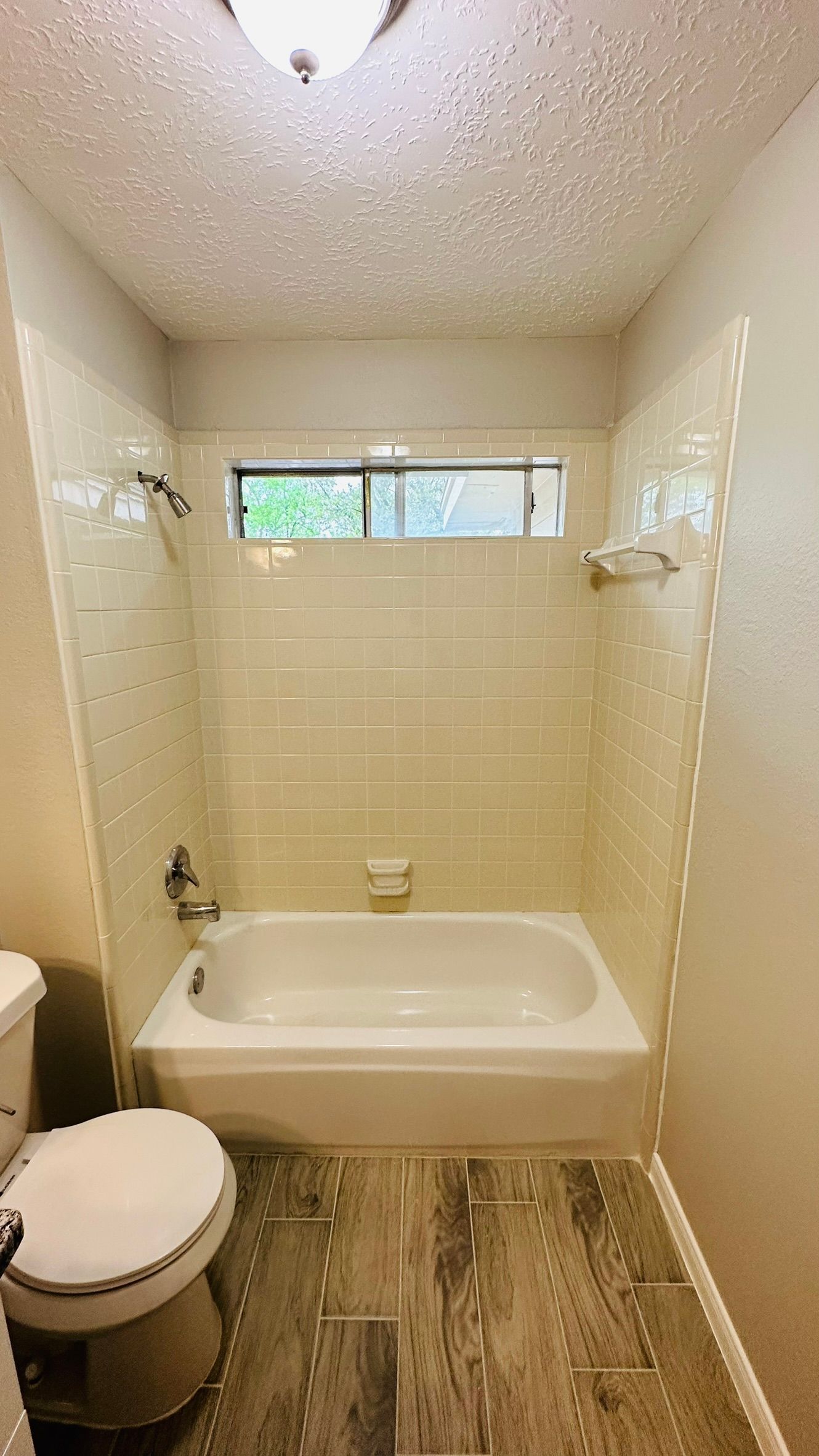 Small bathroom with a white bathtub and beige tiled walls, a window, and wood-look floor.