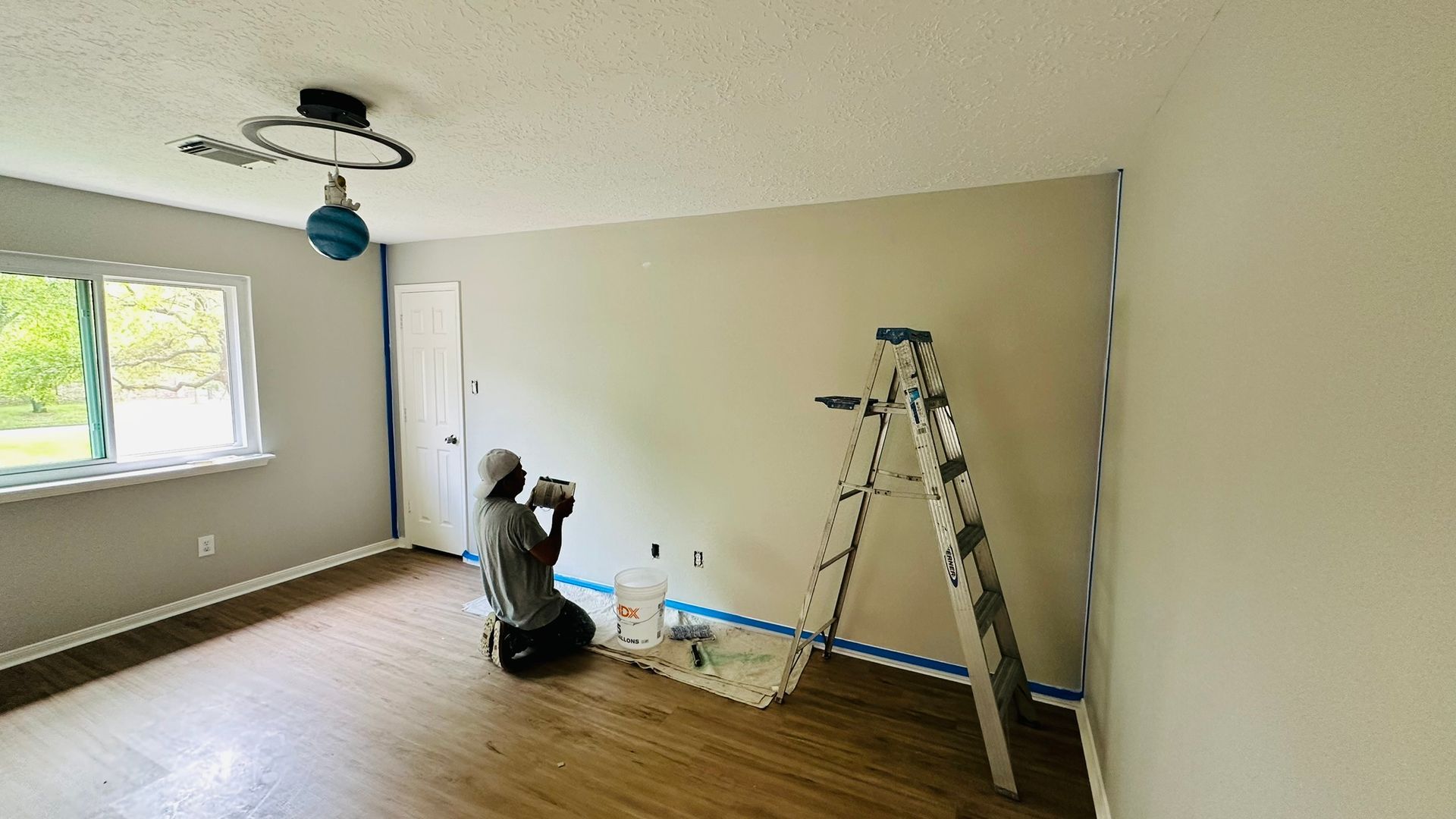 Person painting a room, kneeling with a paint sprayer. A ladder, tape, and paint supplies are nearby.