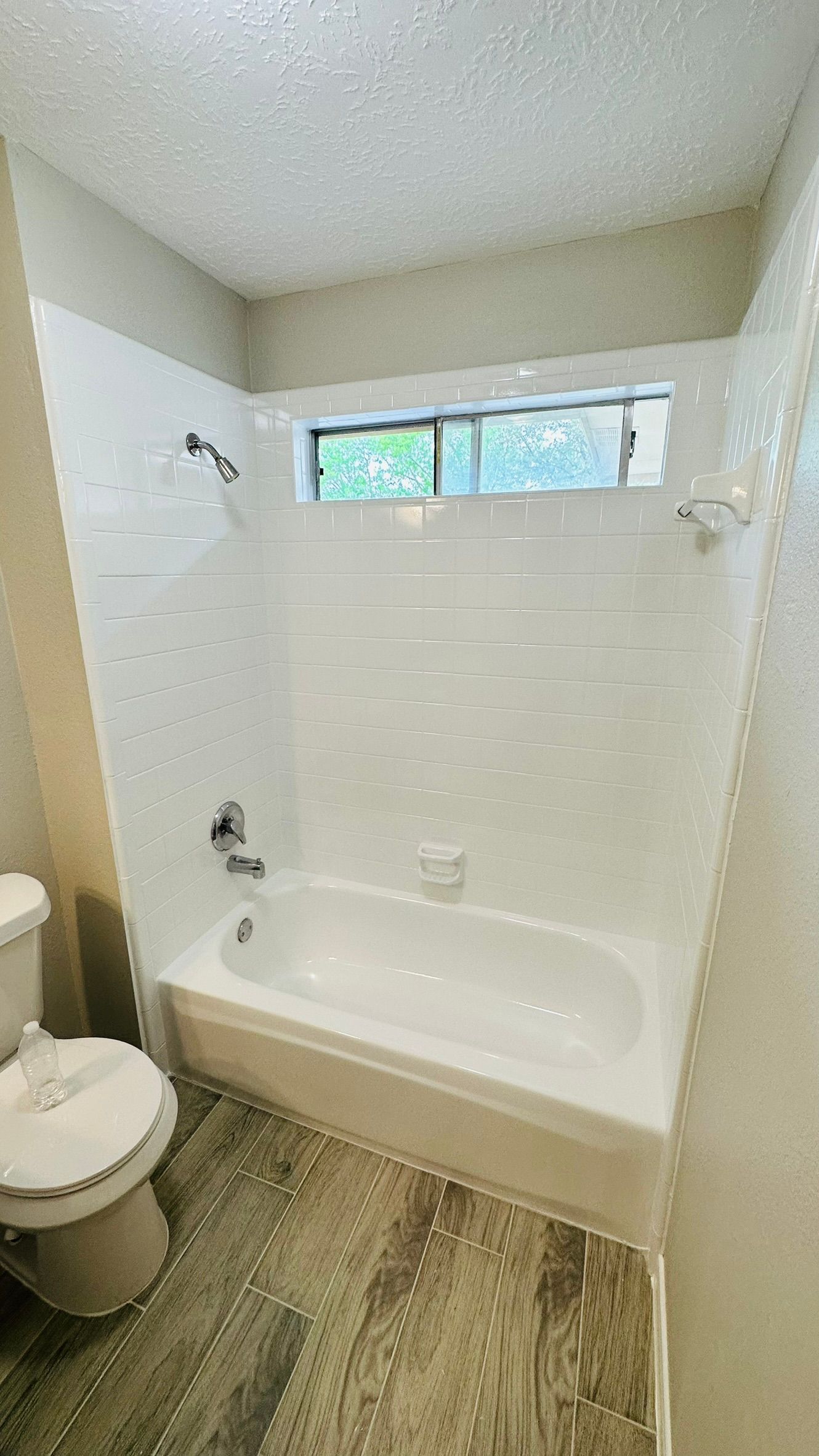 Bathroom with white tub, showerhead, and window above. Light wood-look flooring and a toilet are visible.