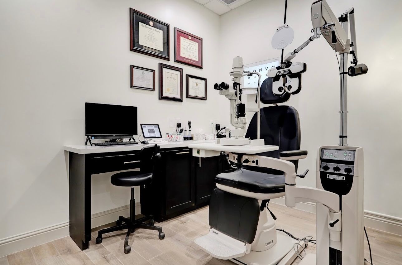 Optometrist's examination room with examination chair, instruments, computer, desk, and framed certificates on the wall.