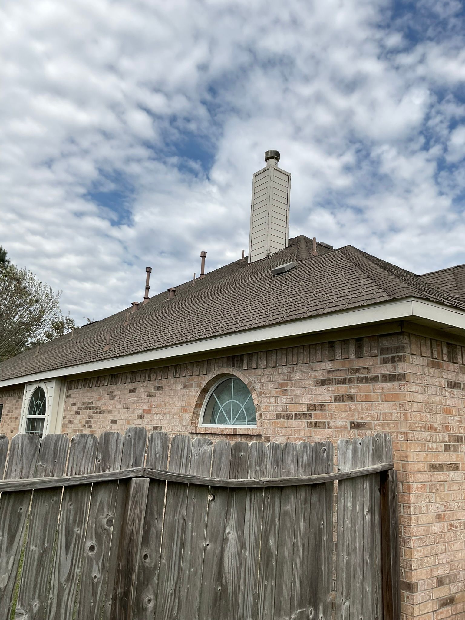 Brick house with a chimney, arched window, and a wooden fence under a cloudy sky.