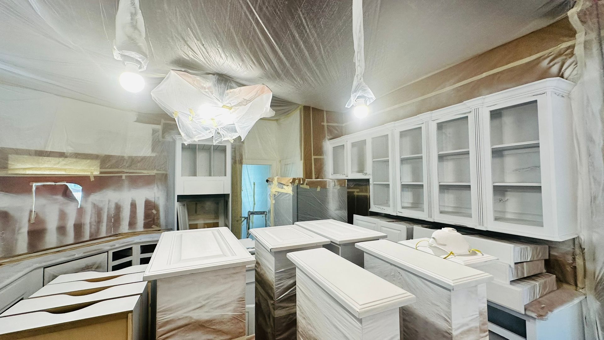 Kitchen being renovated; cabinets and countertops covered in white paint, room draped in plastic.