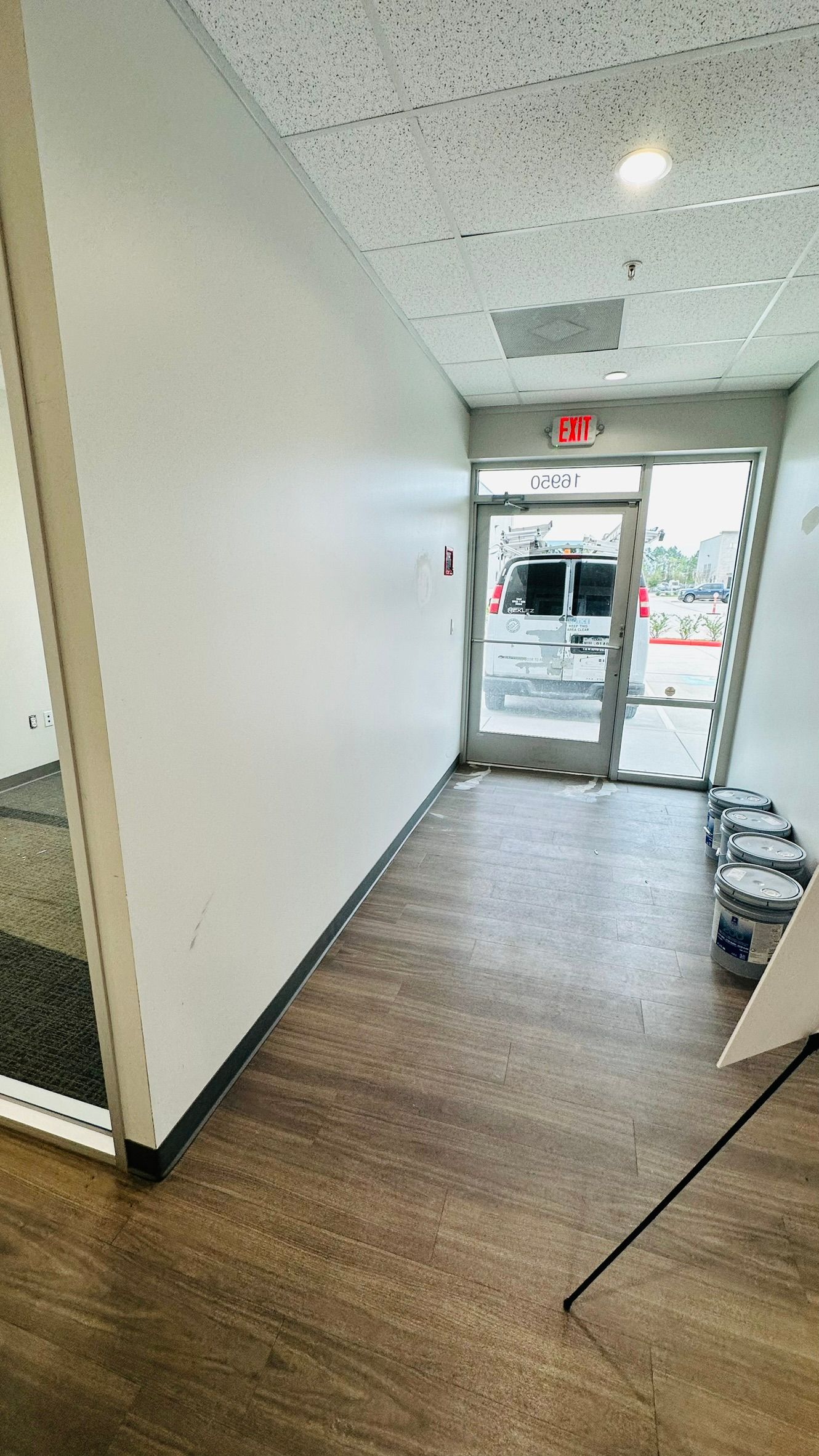 A hallway with white walls and wooden floor leading to an exit door. Painting supplies are visible.
