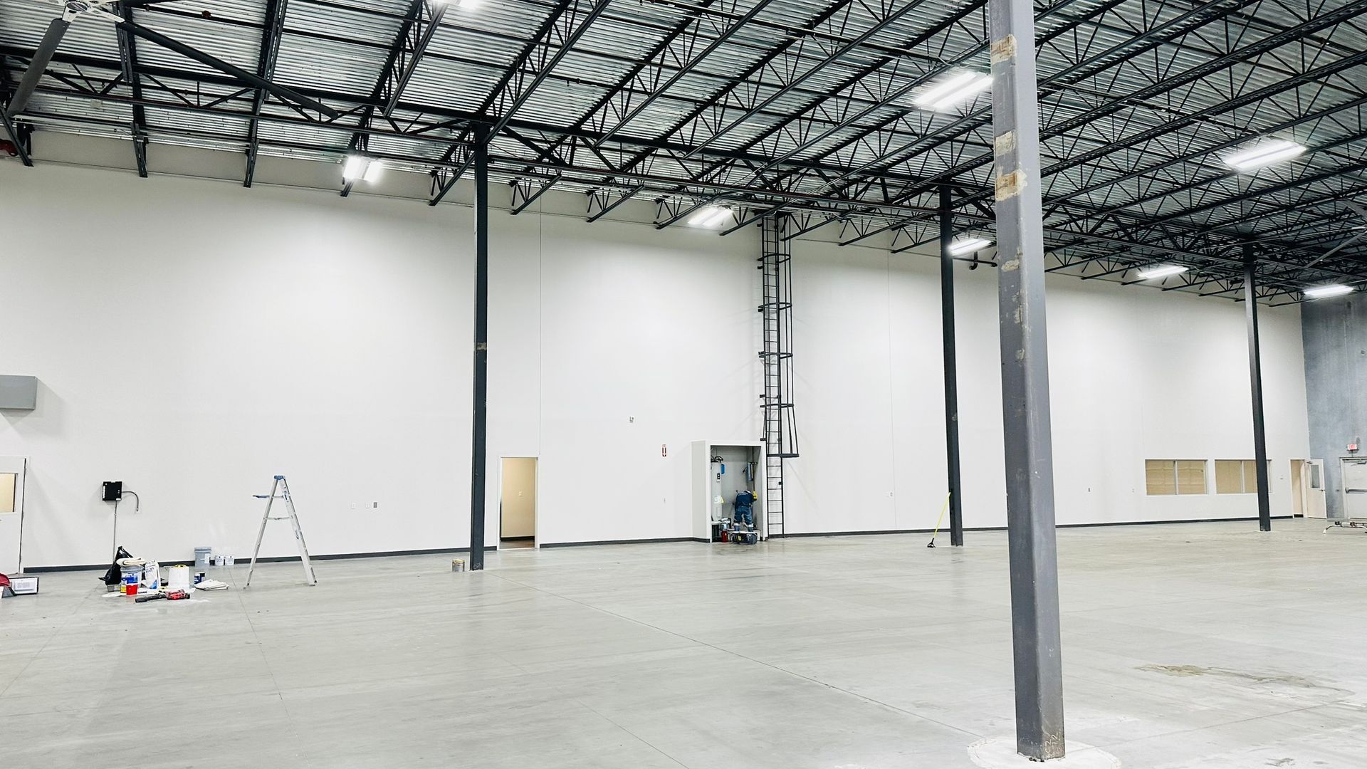 Large empty warehouse interior, white walls, gray floor, black support beams, painting equipment visible.