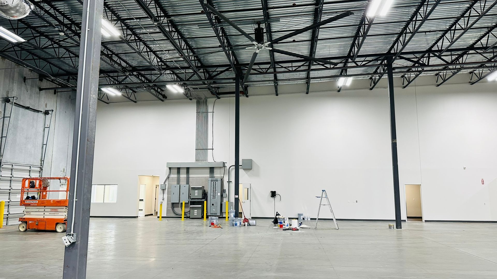 Empty warehouse interior with high ceilings, exposed beams, concrete floor, and equipment.