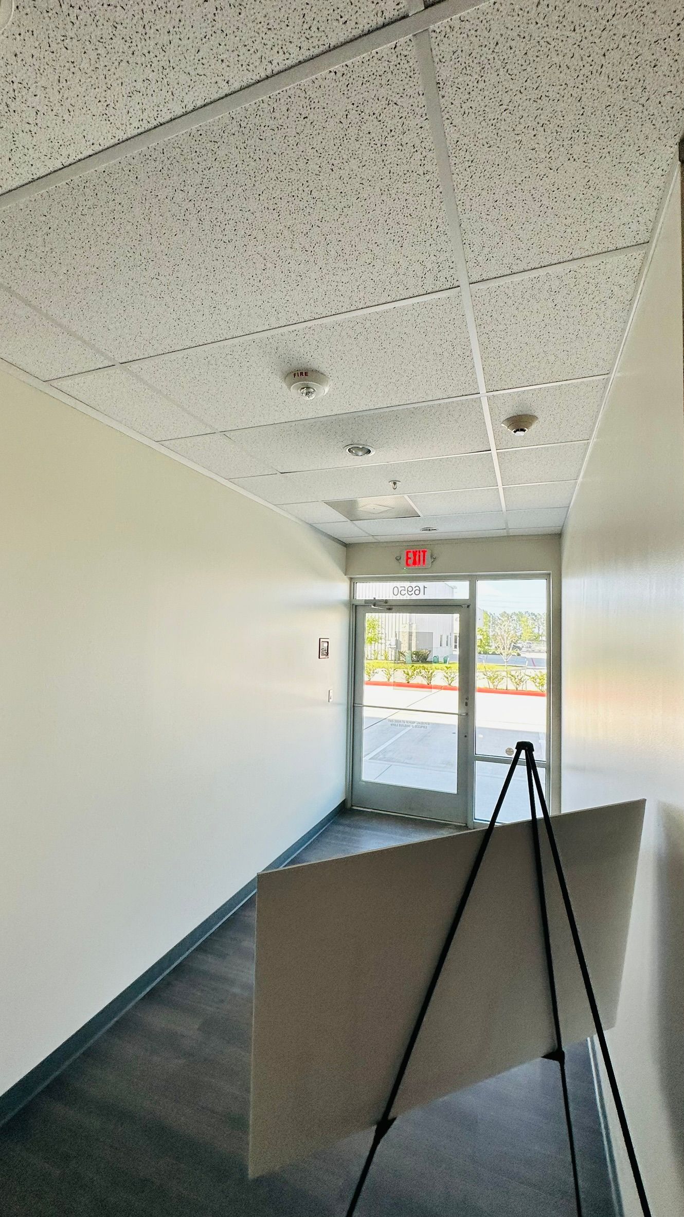 Narrow hallway with white walls, drop ceiling, and exit door at the end. An easel is in the foreground.
