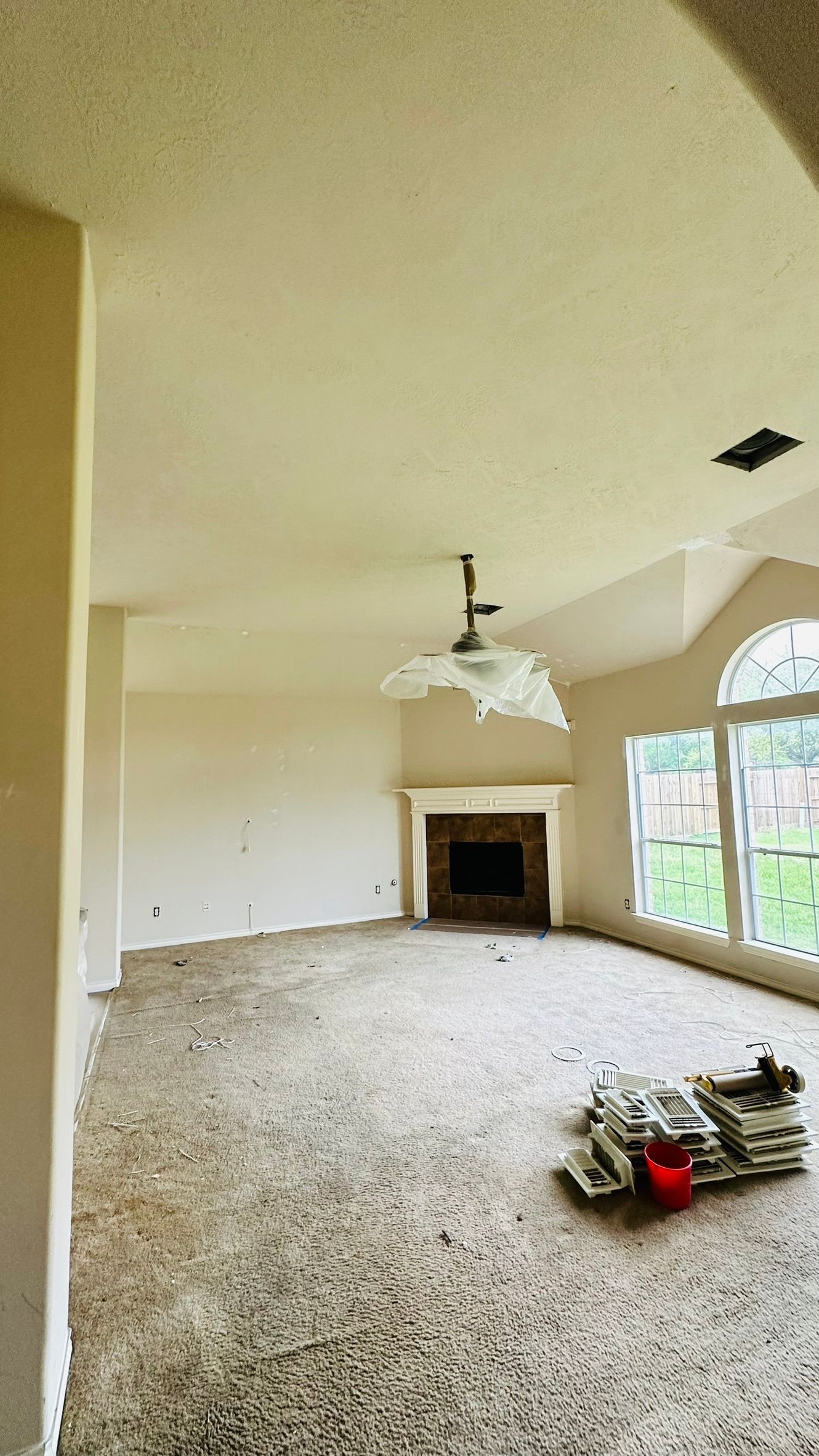 Empty living room, beige walls, fireplace, cracked chandelier, large windows with sunlight.