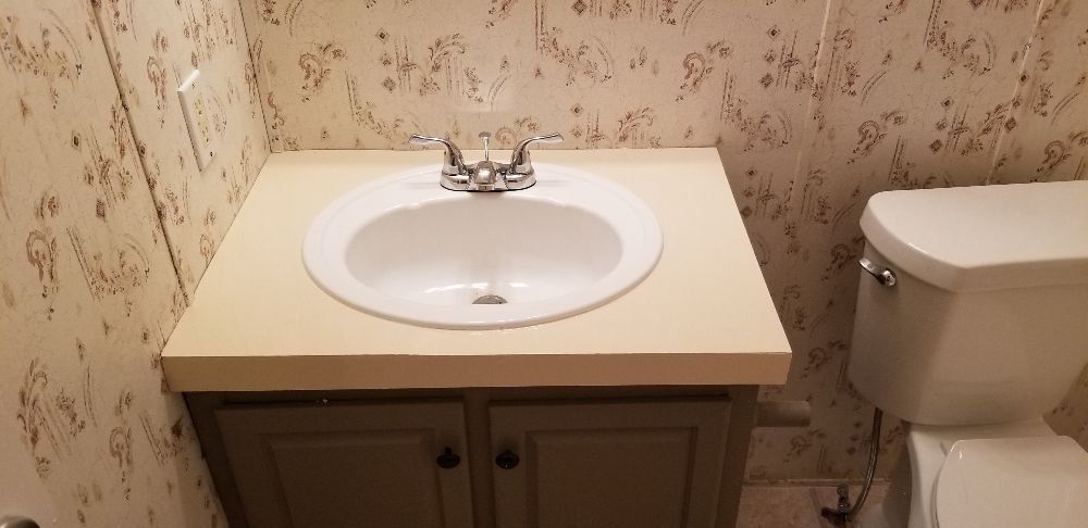 Bathroom vanity with sink and toilet. Beige countertop, light green cabinet, floral wallpaper.