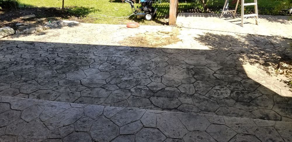 Stone-like stamped concrete patio with cracks, leading to a grassy yard. A lawnmower and ladder visible.
