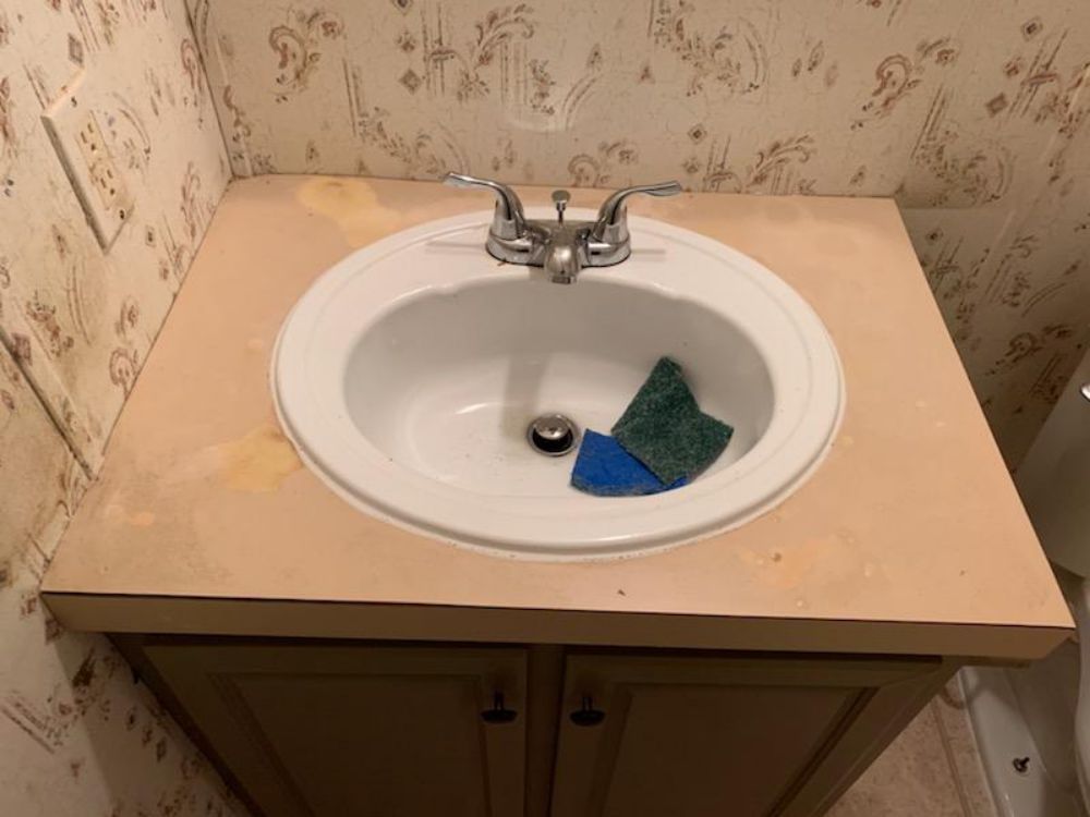 Bathroom sink on a vanity with a white sink, beige countertop, and floral wallpaper. Two sponges rest inside.