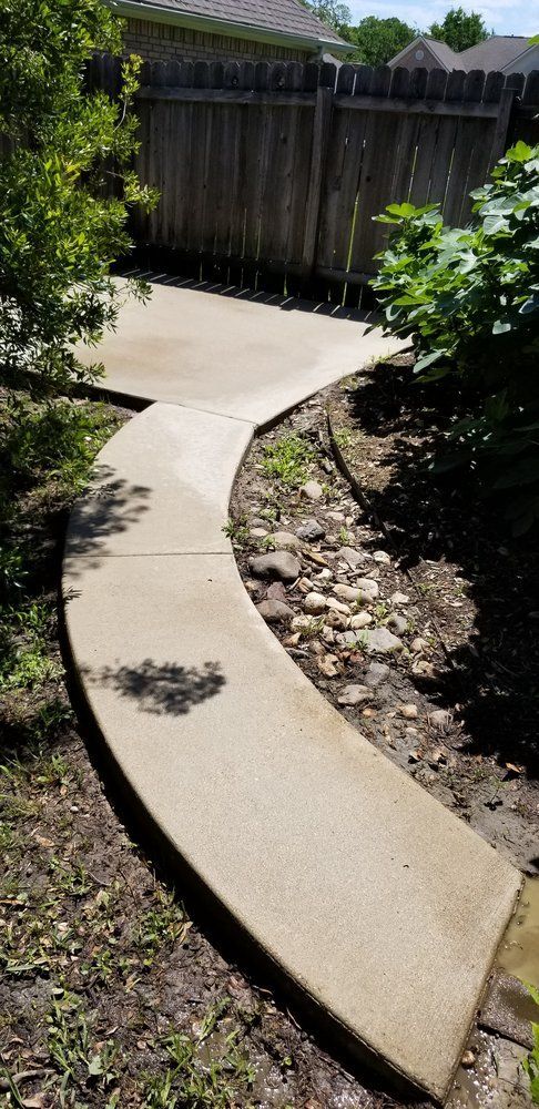 Concrete pathway curving through a yard, bordered by plants and a wooden fence.
