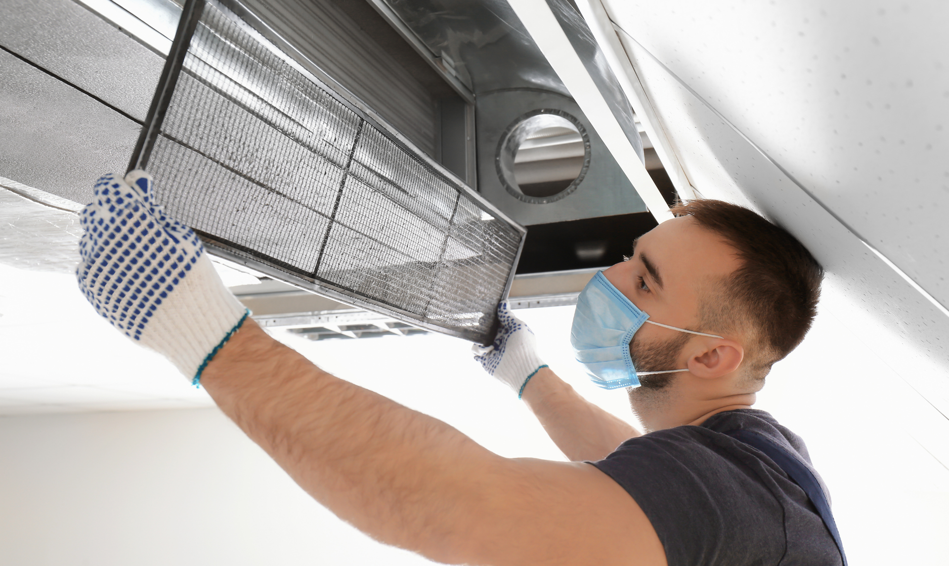 A worker wearing a face mask and gloves removes an air filter from a ceiling-mounted HVAC unit.