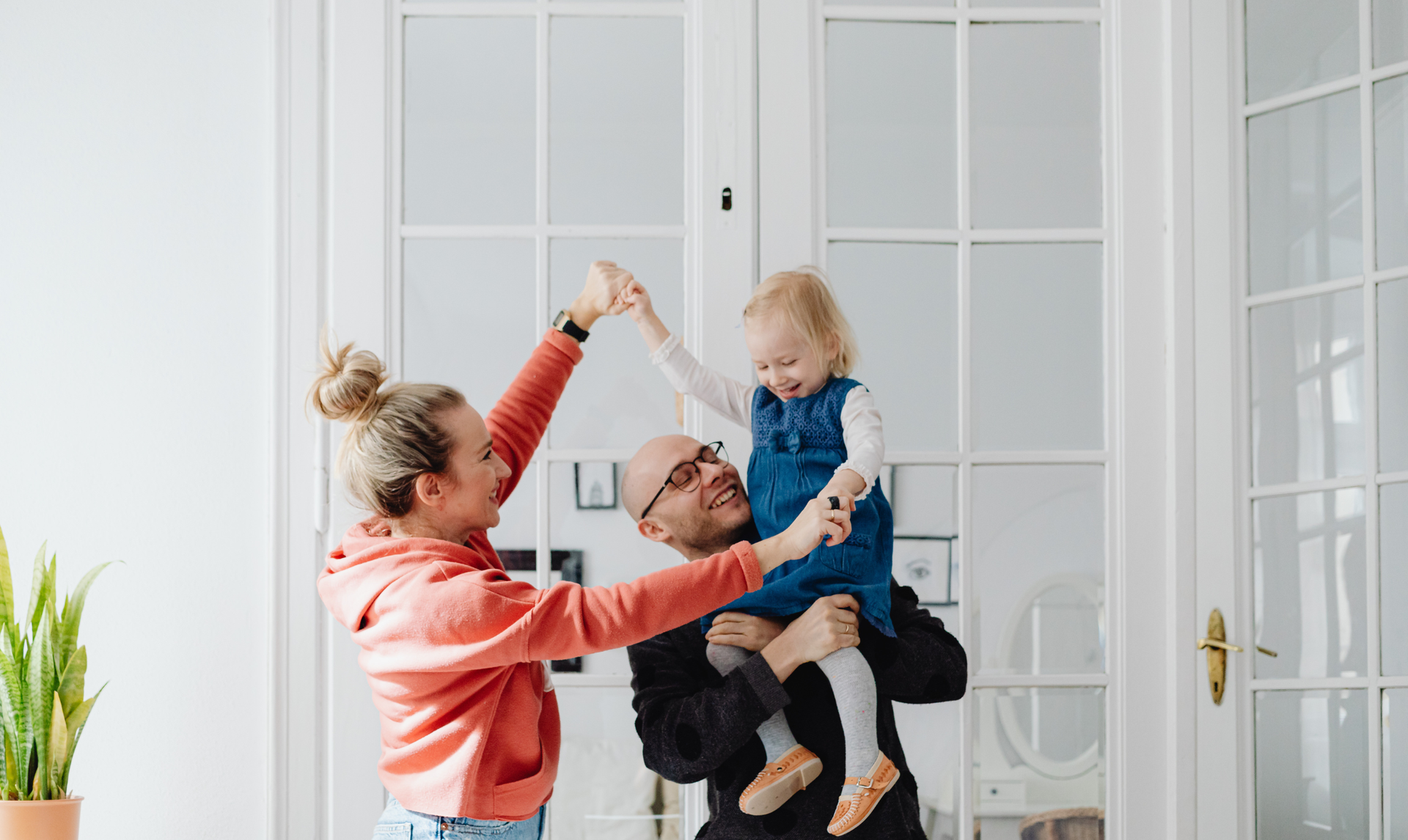 A person in a coral hoodie and a person in a black shirt lifting a child, all laughing in front of a glass-paneled door.