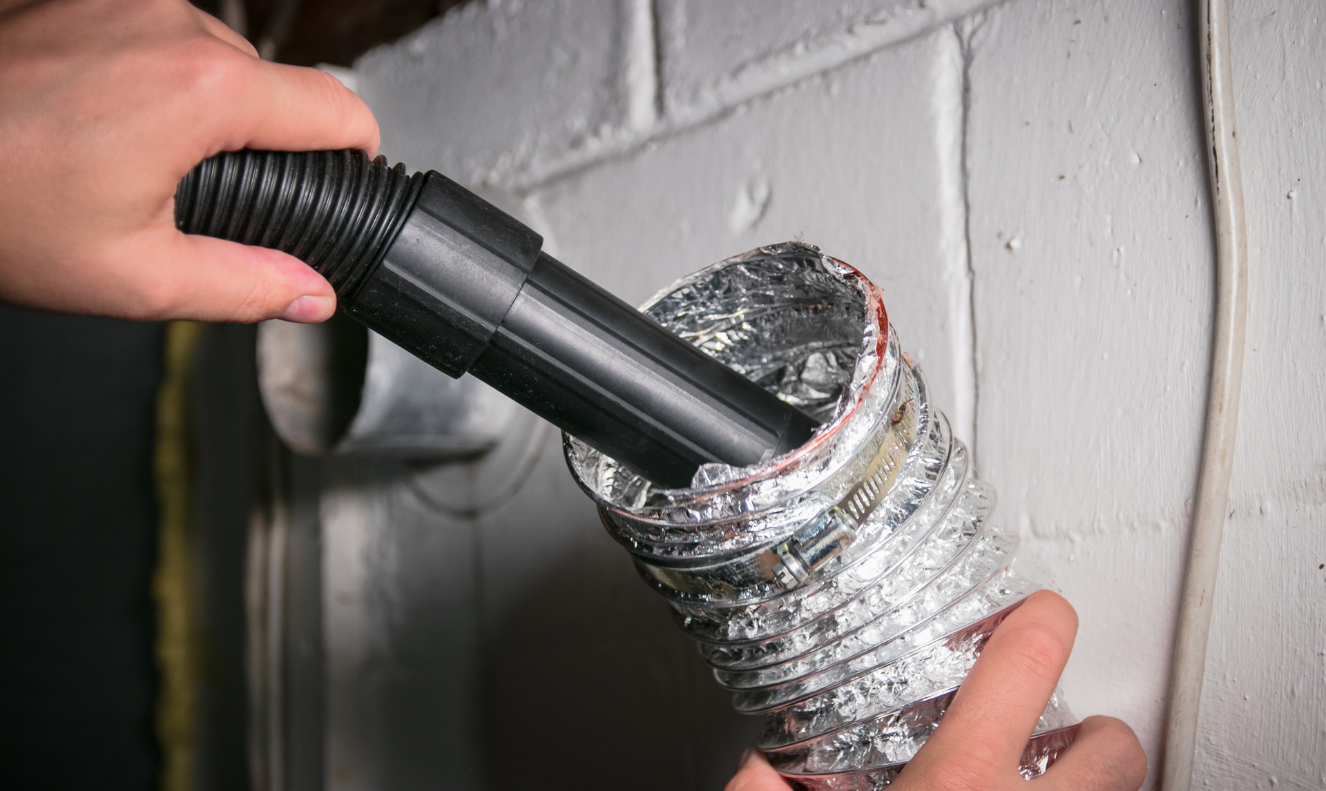 Hands use a vacuum attachment to clean the inside of a flexible silver dryer vent hose mounted on a white brick wall.