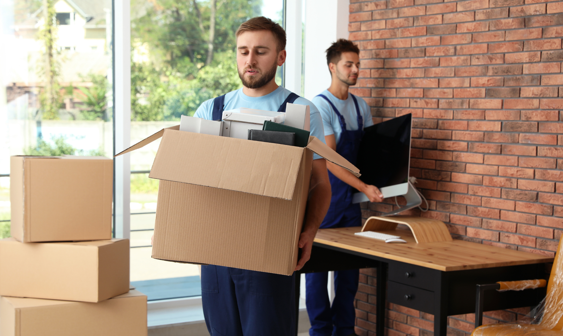 Two workers in blue uniforms move office supplies, one carrying a cardboard box while the other carries a computer monitor.