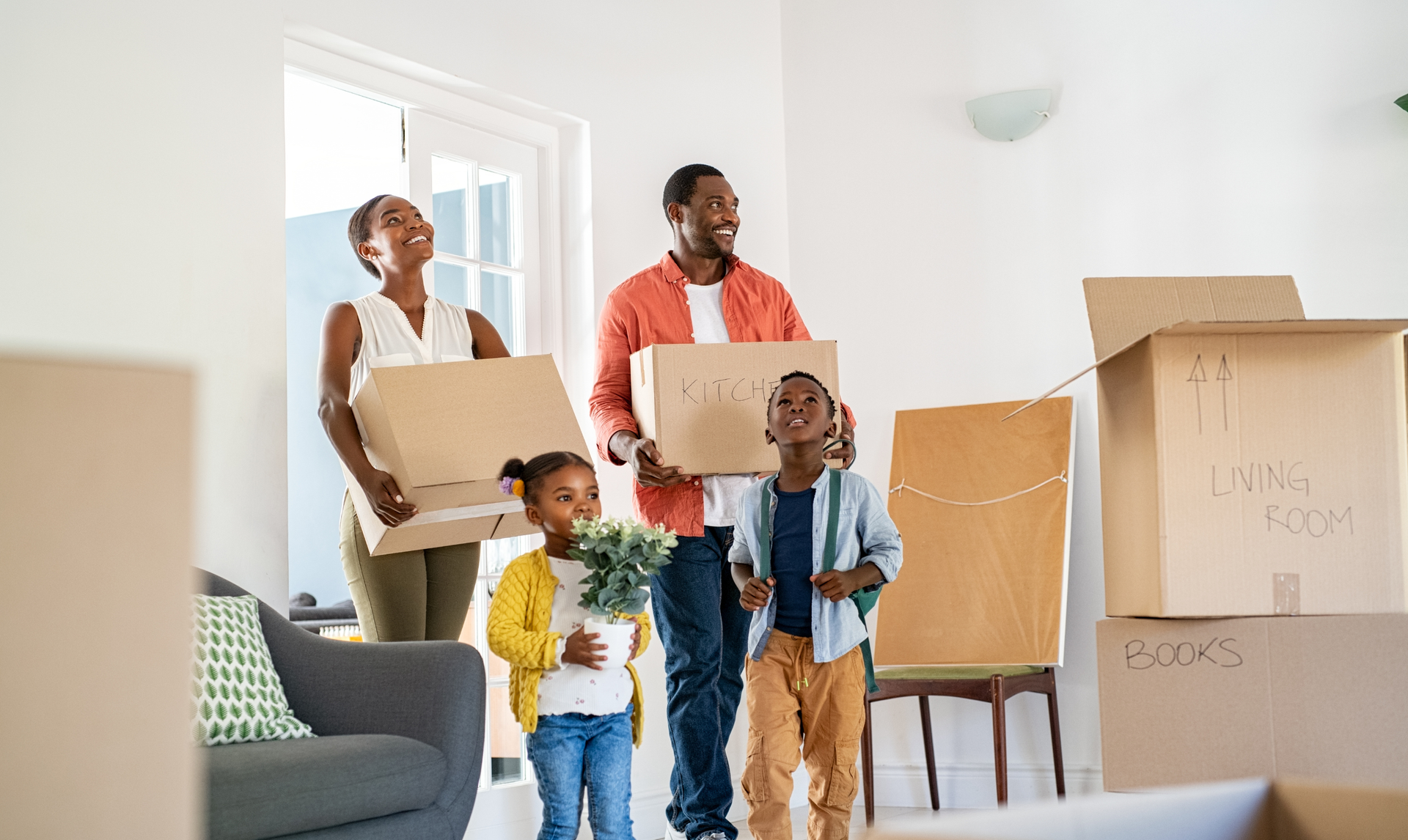 A family stands in their new home holding moving boxes, looking up with smiles while surrounded by packing supplies.