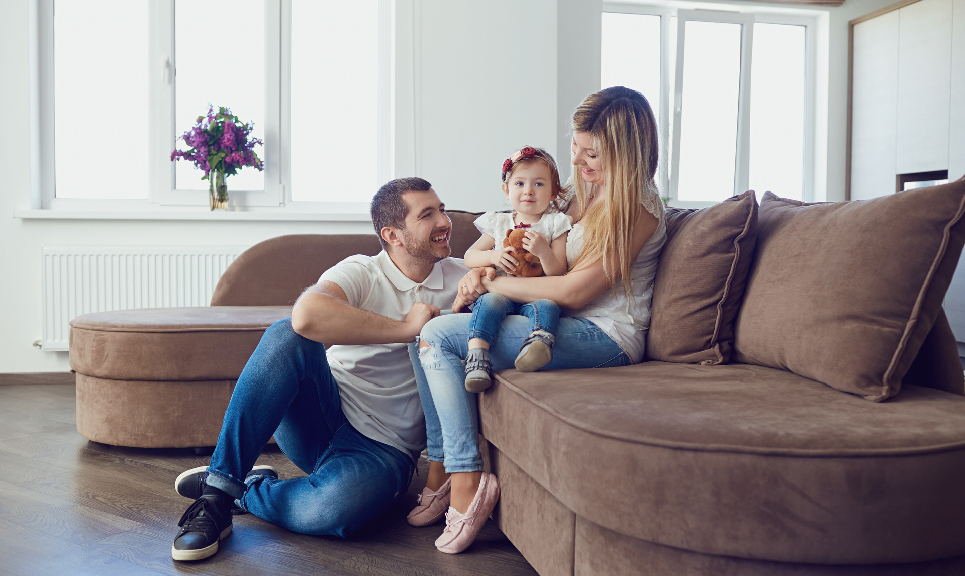 A small family sits together on a large brown sofa in a bright, modern living room.