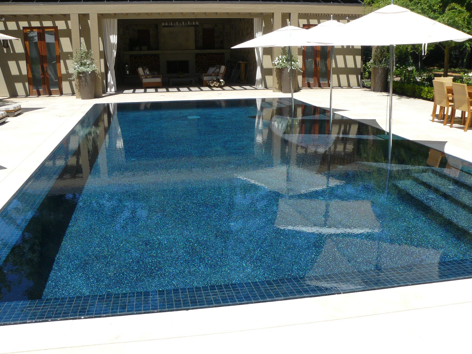 Outdoor swimming pool with blue water, white umbrellas, and lounge chairs beside a patio.