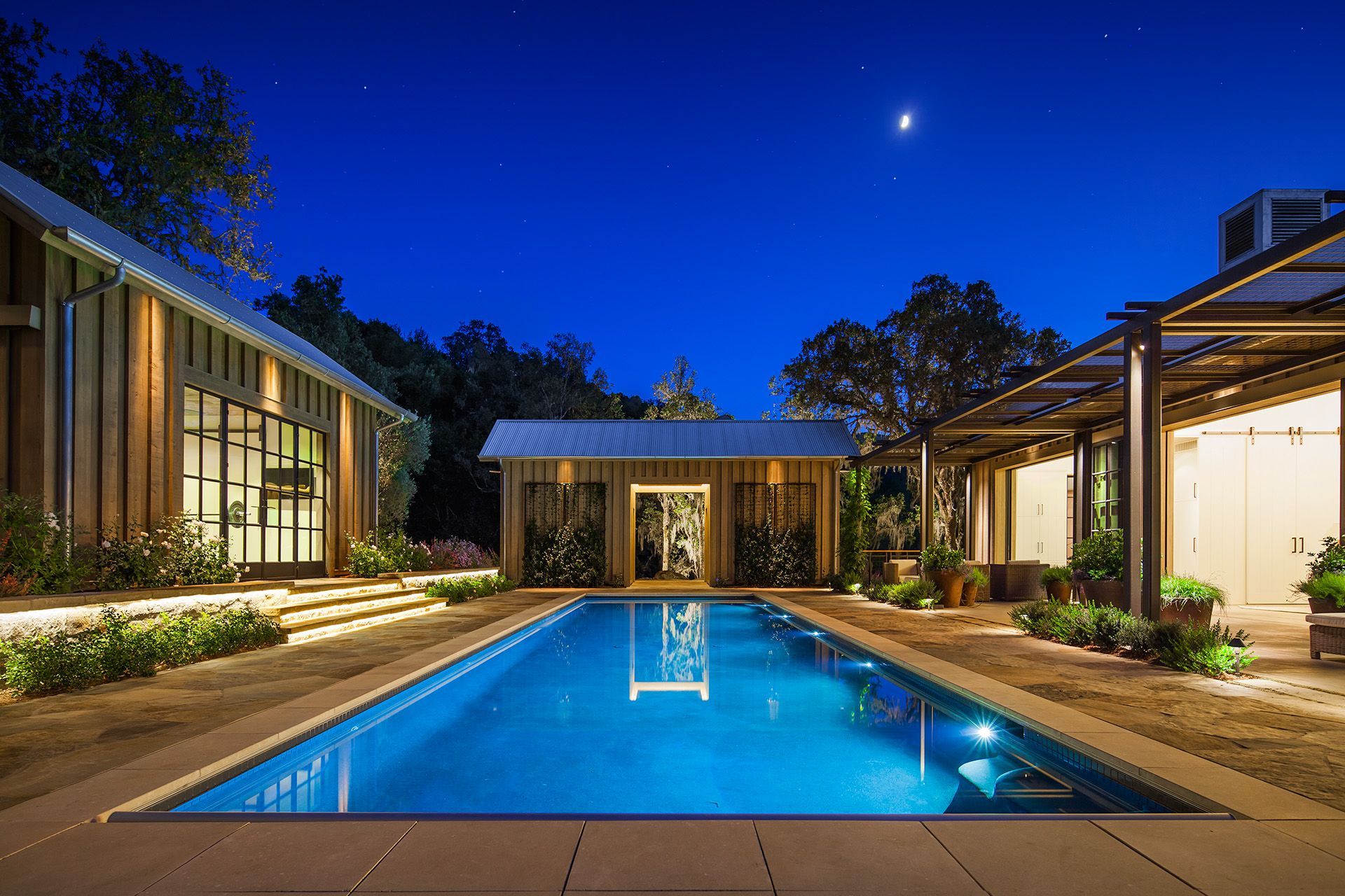 Modern home patio at night with a lit rectangular pool, surrounding glass buildings, and starry sky