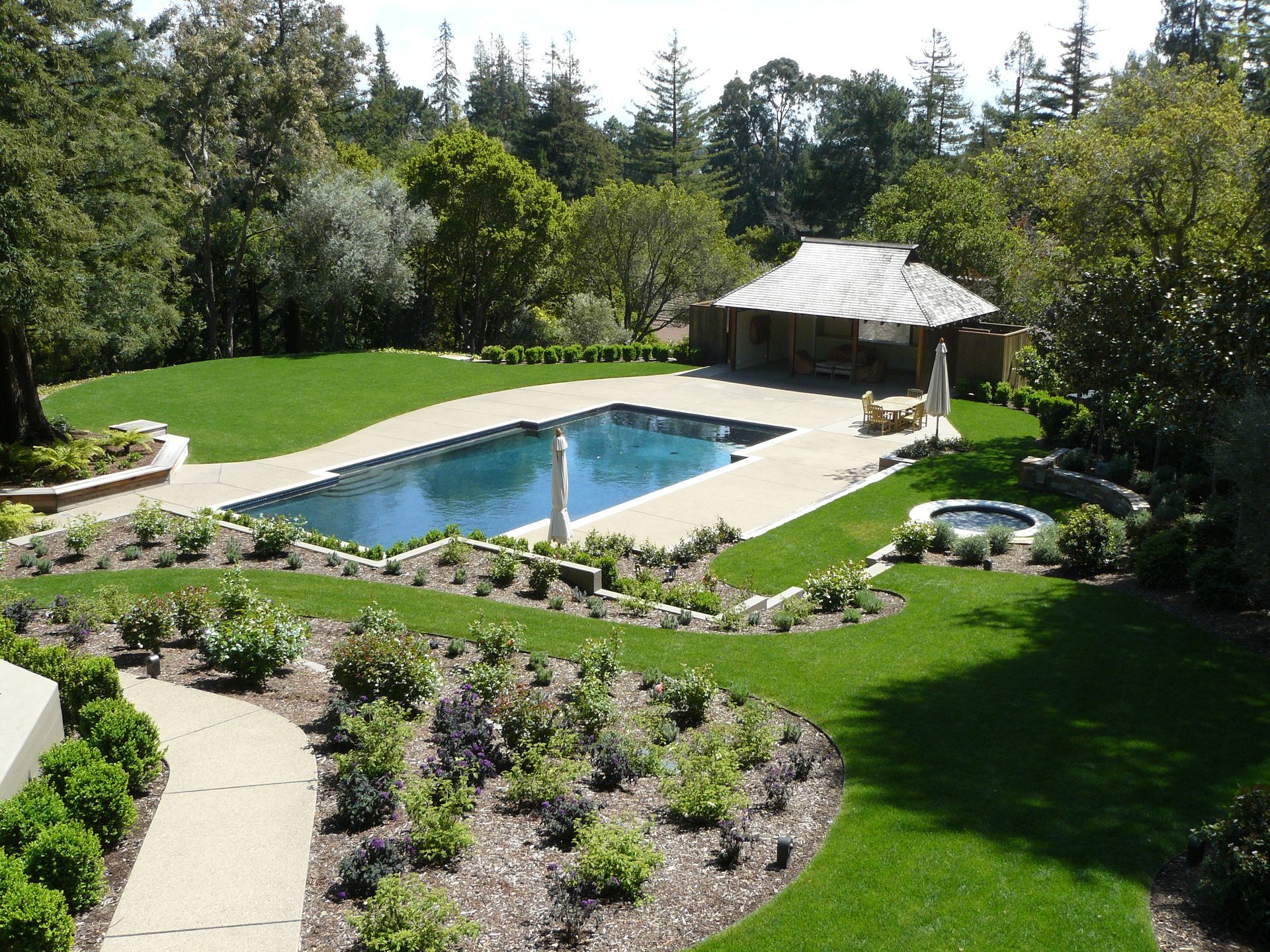 Backyard with a swimming pool, green lawn, garden beds, and a covered patio surrounded by trees