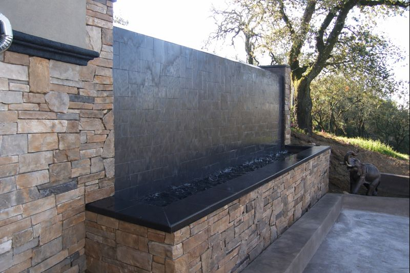 Stone outdoor water feature with a black wall and basin beside a paved patio and trees