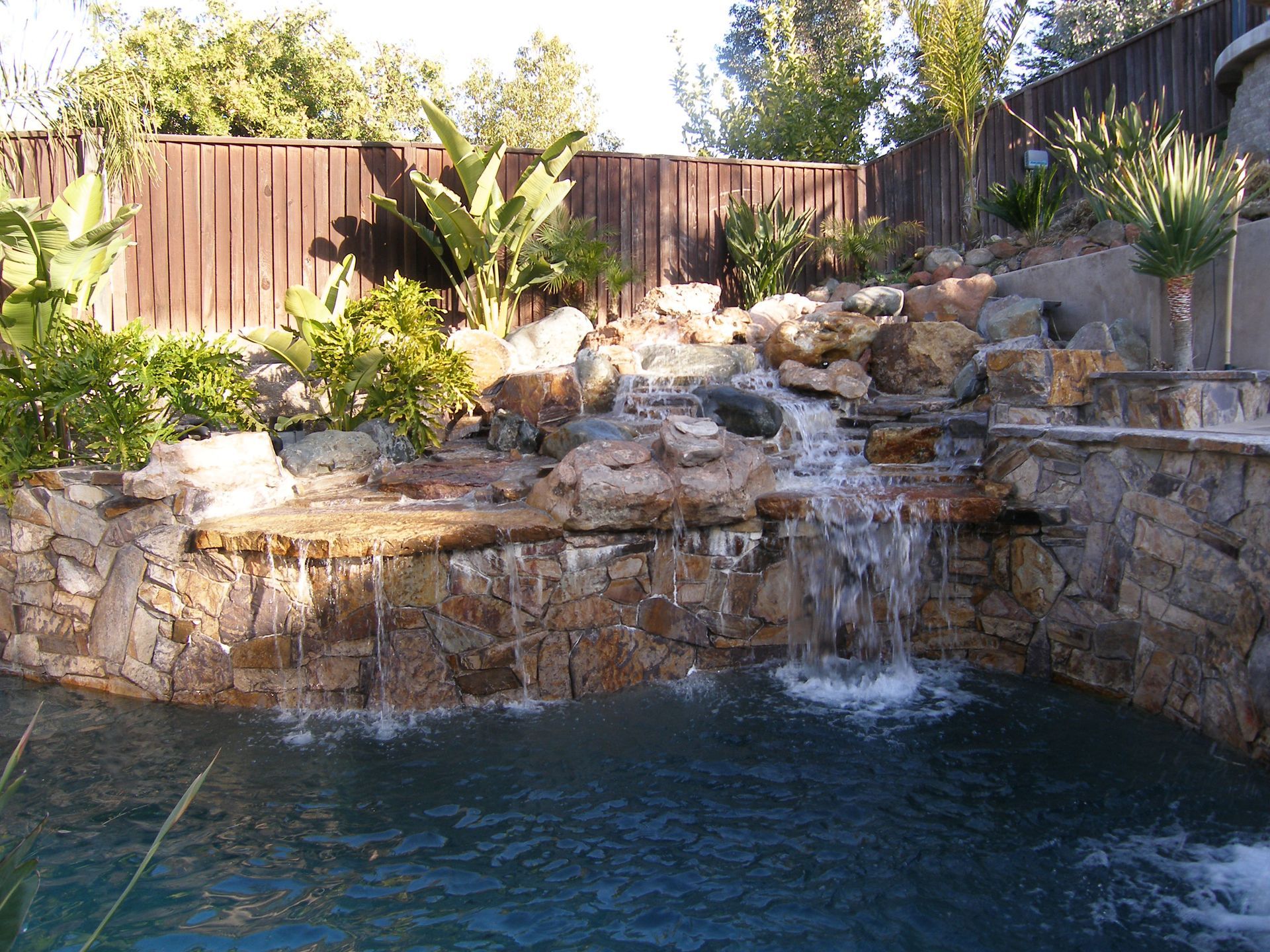 Backyard rock waterfall cascading into a blue pool, surrounded by plants and a wooden fence.