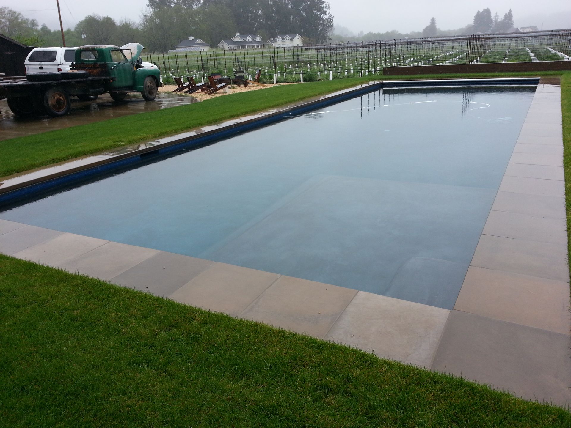 Large rectangular swimming pool with still water, bordered by grass and paving, with trucks in the background