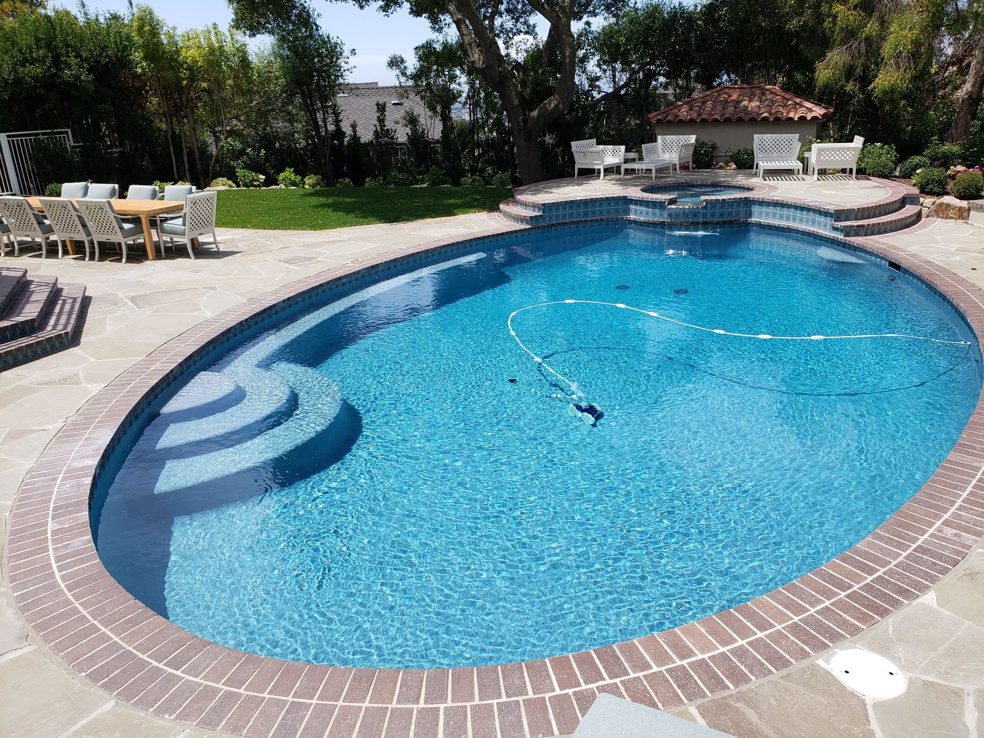 Backyard swimming pool with blue water, brick edge, lounge chairs, and patio furniture under trees