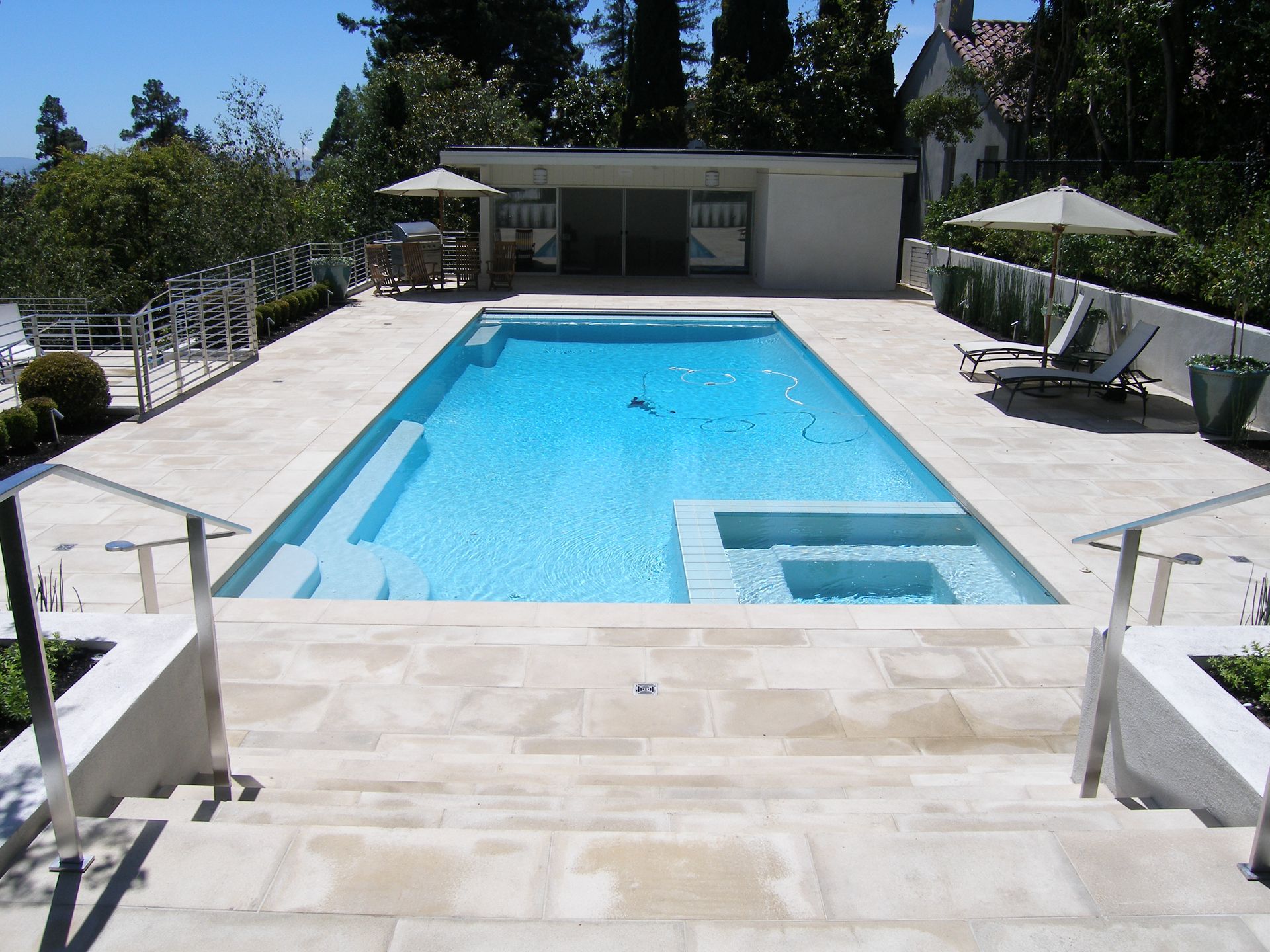 Bright backyard pool with white stone deck, lounge chairs, and a small pool house in the background