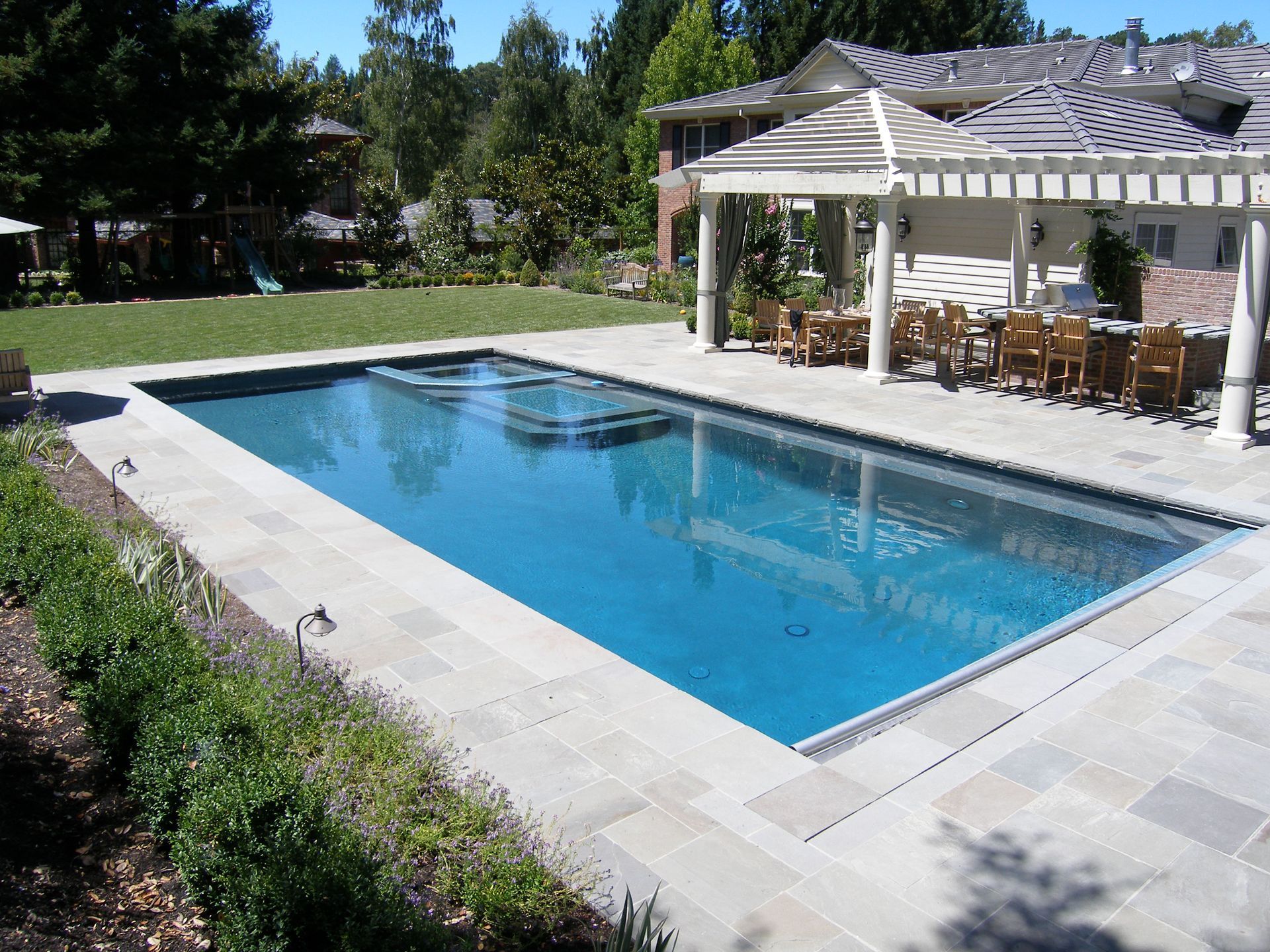 Rectangular blue swimming pool beside a white pergola with patio furniture in a landscaped yard