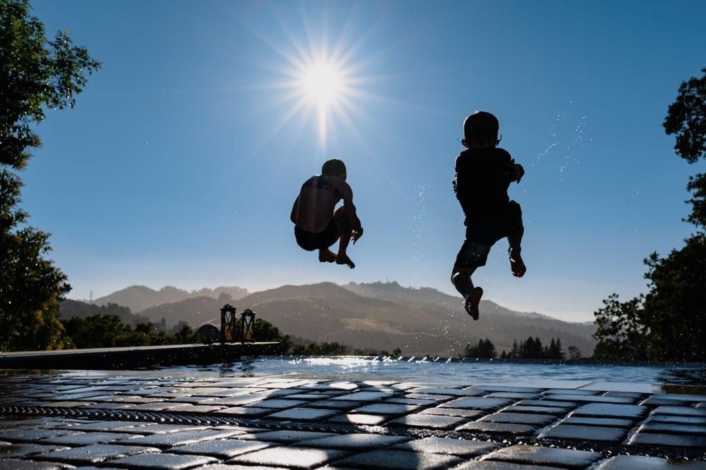 Two silhouetted people jump on a wet outdoor surface under a bright sun, with mountains in the background.