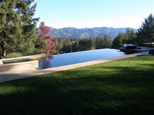 Modern house with an infinity pool overlooking a tree line. Two lounge chairs, a striped umbrella, and blue sky. Modern house with an infinity pool overlooking a tree line. Two lounge chairs, a striped umbrella, and blue sky.